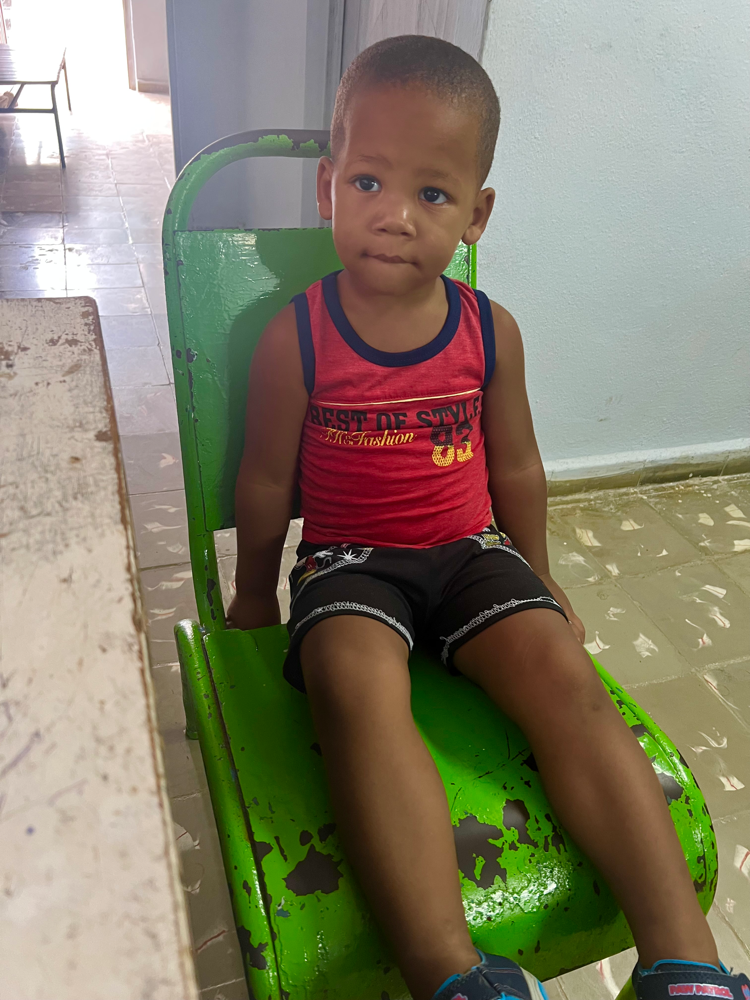 A young boy sitting on a green, distressed metal chair indoors.