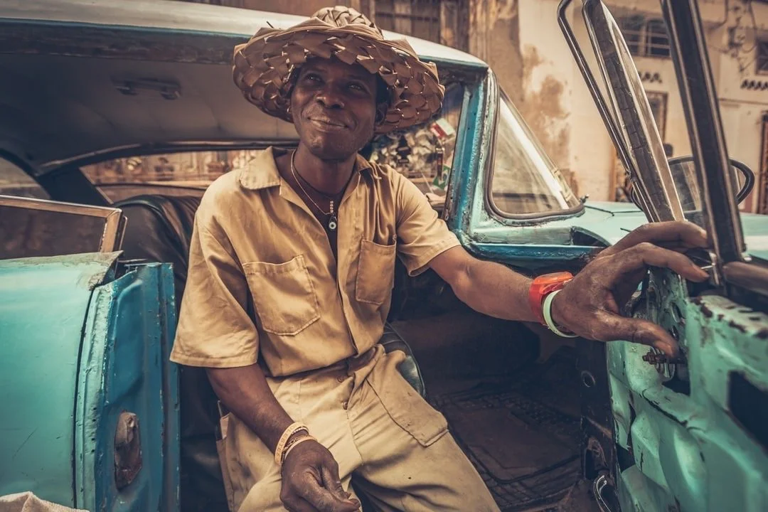 A man wearing a straw hat and tan clothes sitting in an old, weathered vintage vehicle. He appears to be smiling and holding the glove compartment.
