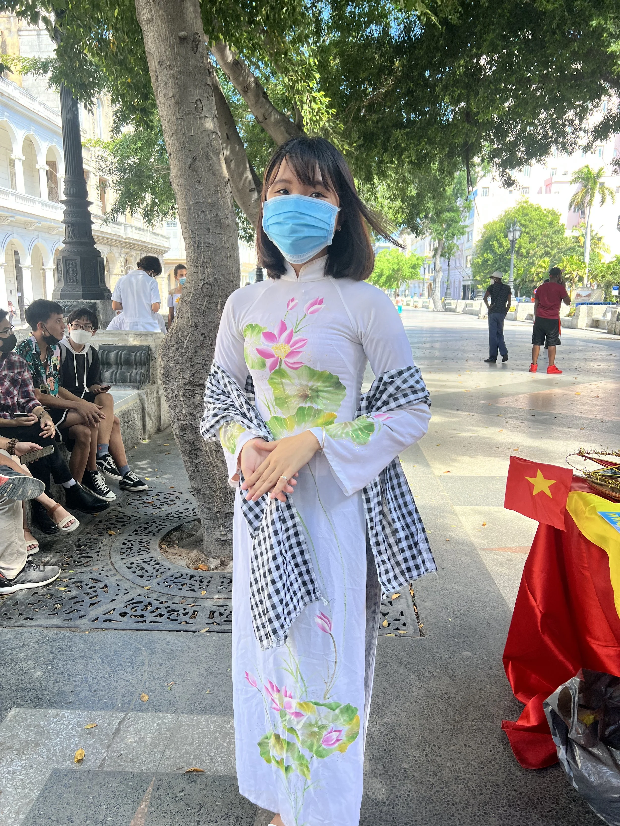 A woman wearing a blue face mask, traditional dress with lotus flower designs, stands outdoors near a tree, with people seated on benches and walking in the background.