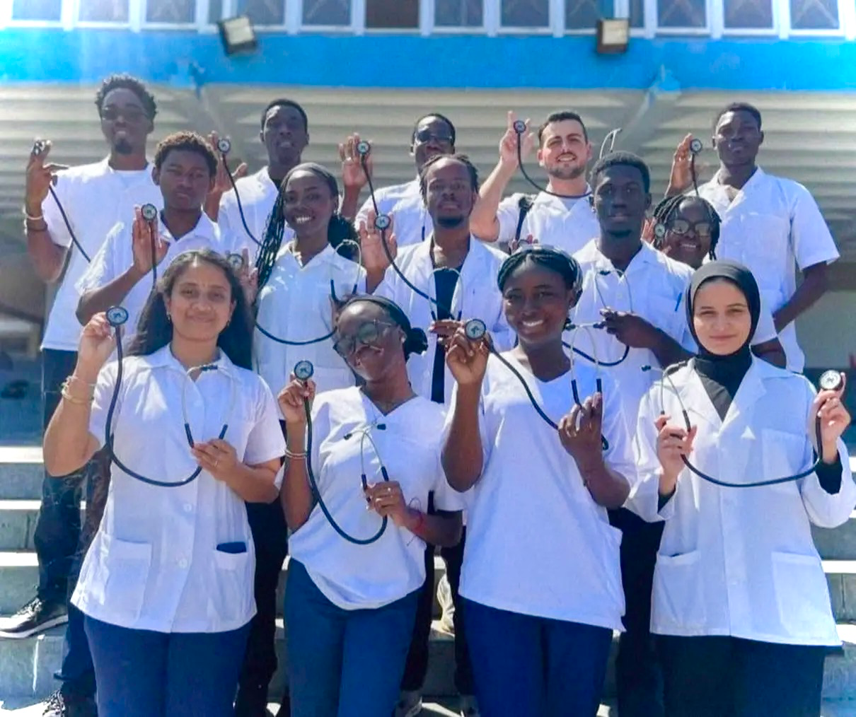 Group of healthcare professionals in white coats and scrubs, holding stethoscopes, smiling outdoors in front of a building.