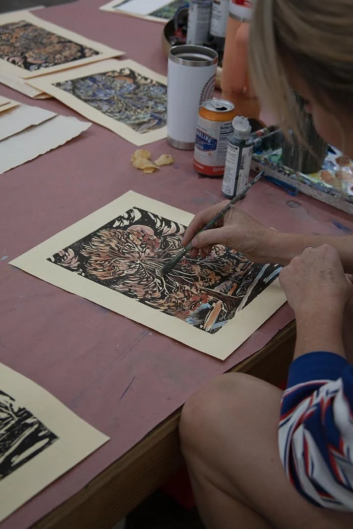 Michelle Blade hand painting wood block prints using paint and brushes on paper, with art supplies and printed pictures on the table.