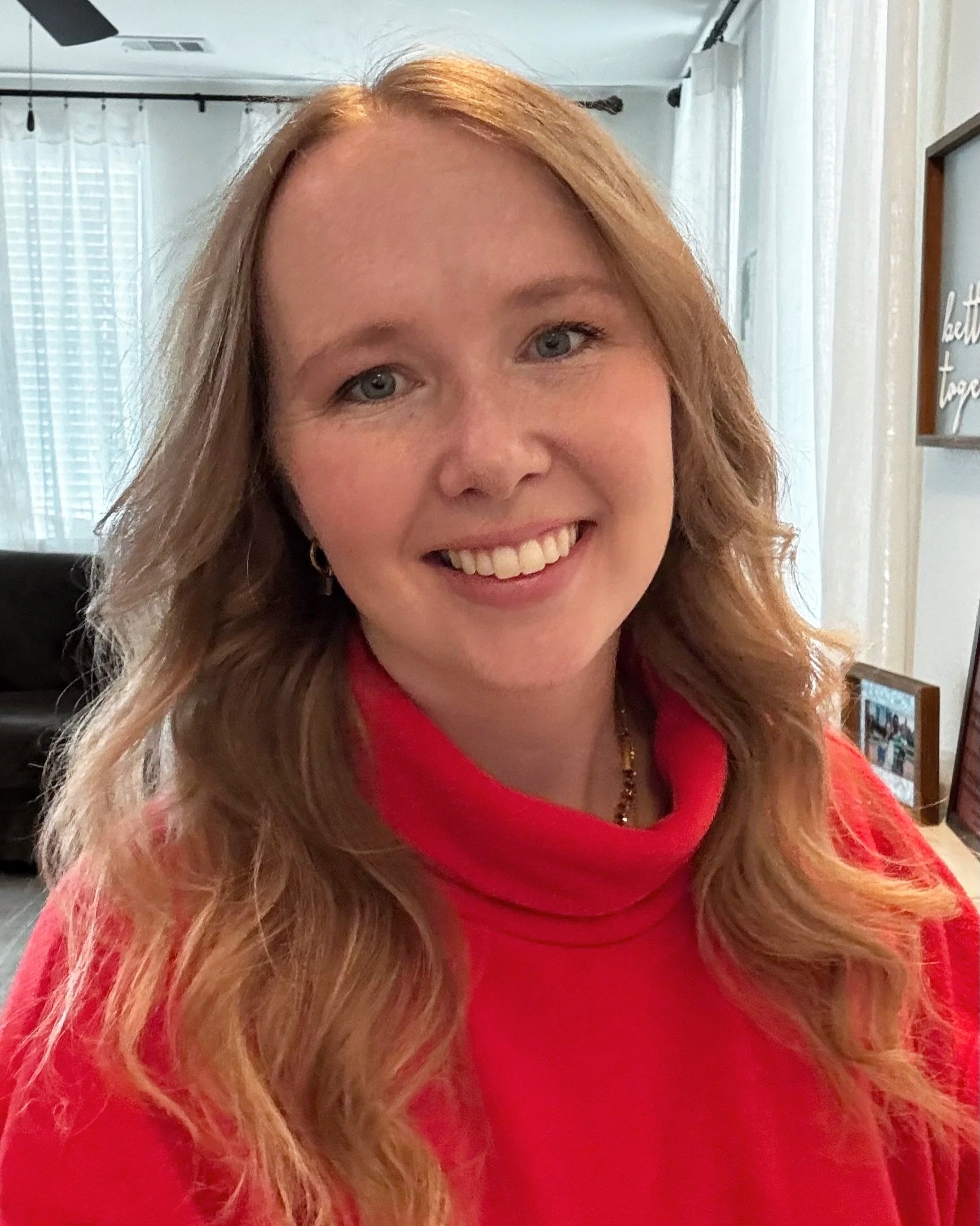 A woman with long wavy red hair, blue eyes, and fair skin is smiling at the camera. She is wearing a red turtleneck sweater and gold earrings, standing in a bright living room with sheer white curtains and decorative framed artwork on the wall.