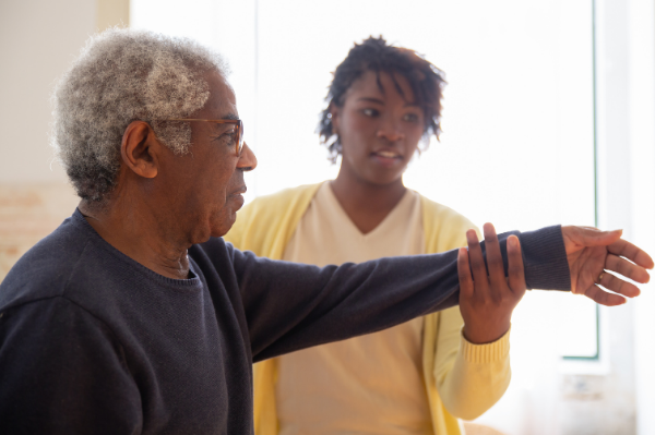 An elderly woman with gray hair and glasses stretches her arm while a young woman with short curly hair observes in a well-lit room.