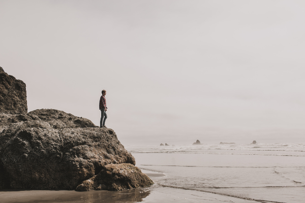 A person standing on a large rock at the beach, looking out over the ocean on a cloudy day.