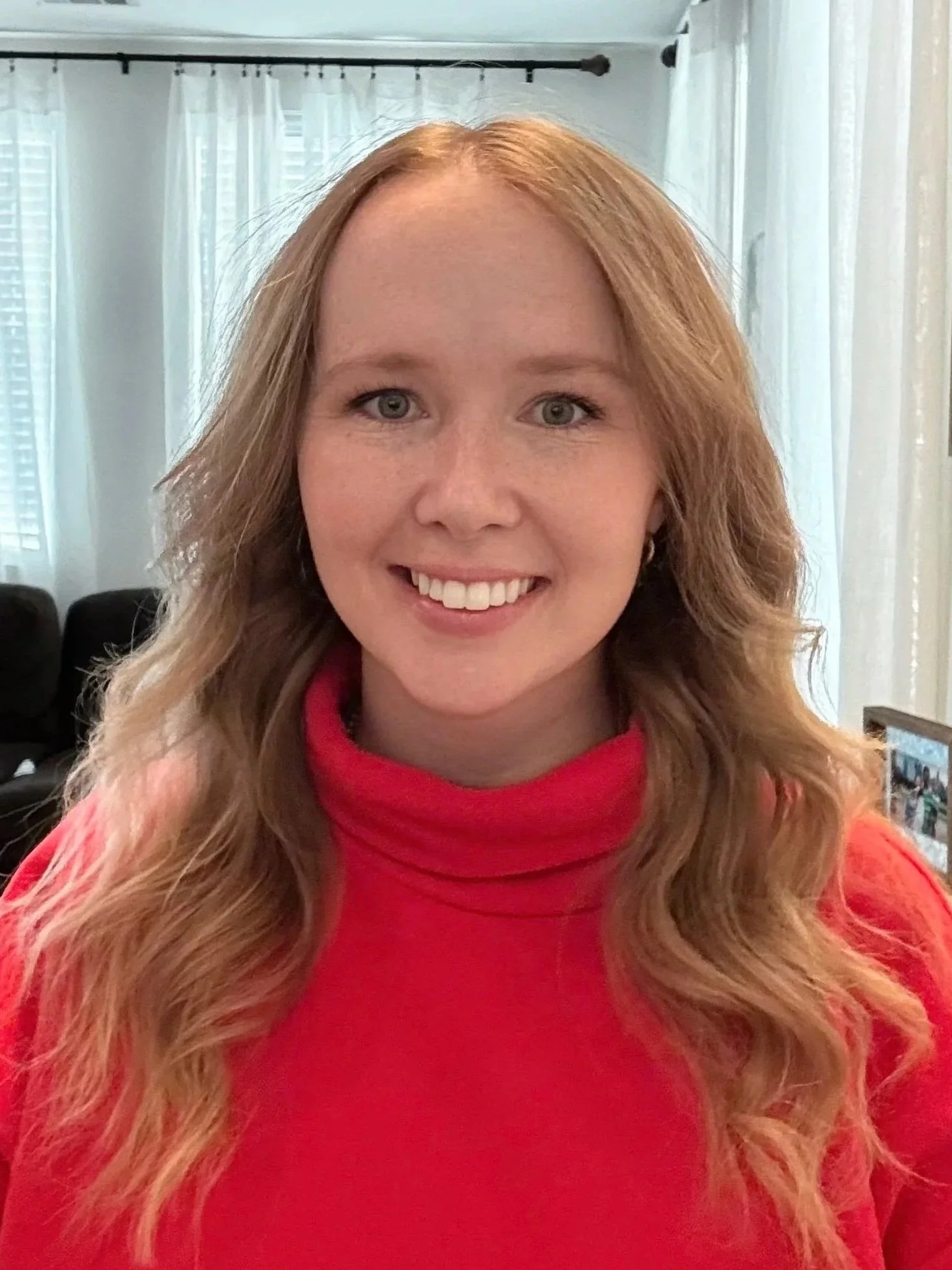A woman with long, wavy red hair wearing a red turtleneck sweater, smiling indoors with white curtains and windows in the background.