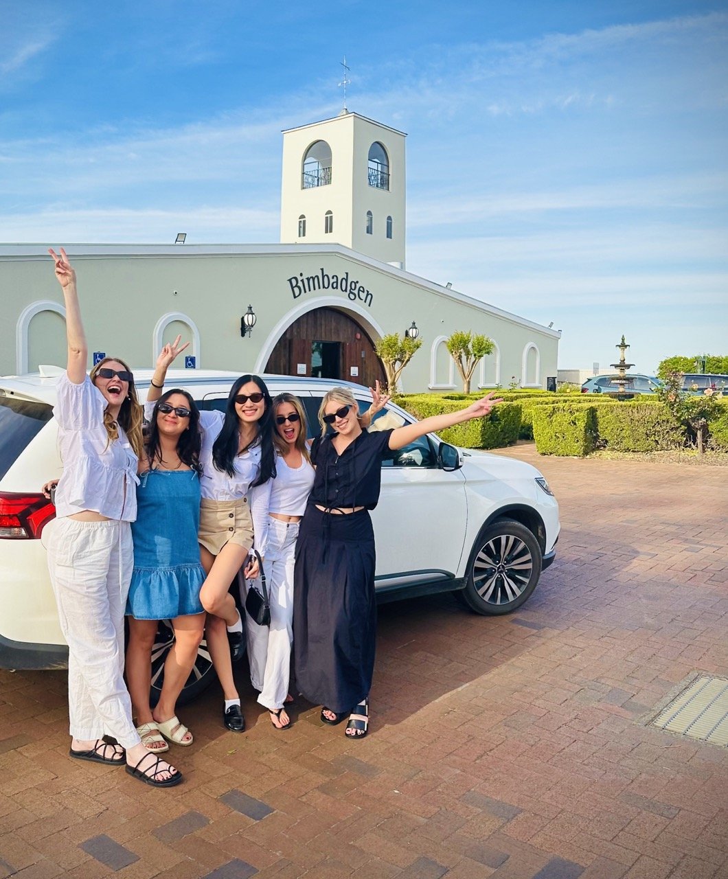 Five women smiling and posing in front of a white vehicle outside a building with the sign 'Bimbadagen', under a clear blue sky.