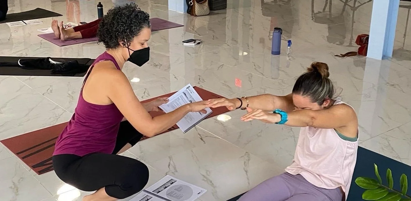 A woman in a purple tank top and black face mask is assisting another woman with stretching exercises in a gym or fitness studio. The woman being assisted is seated on a chair, wearing a light pink tank top, and is reaching forward with both arms.