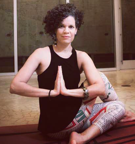 A woman practicing yoga indoors, sitting cross-legged on a mat with hands pressed together in a prayer position.