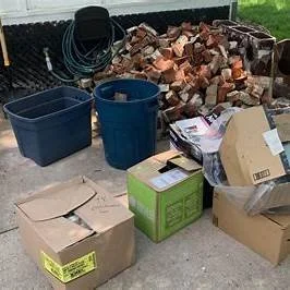 A pile of cardboard boxes, two blue plastic storage containers, a bundle of black hoses, and a heap of bricks outdoors on a concrete surface.