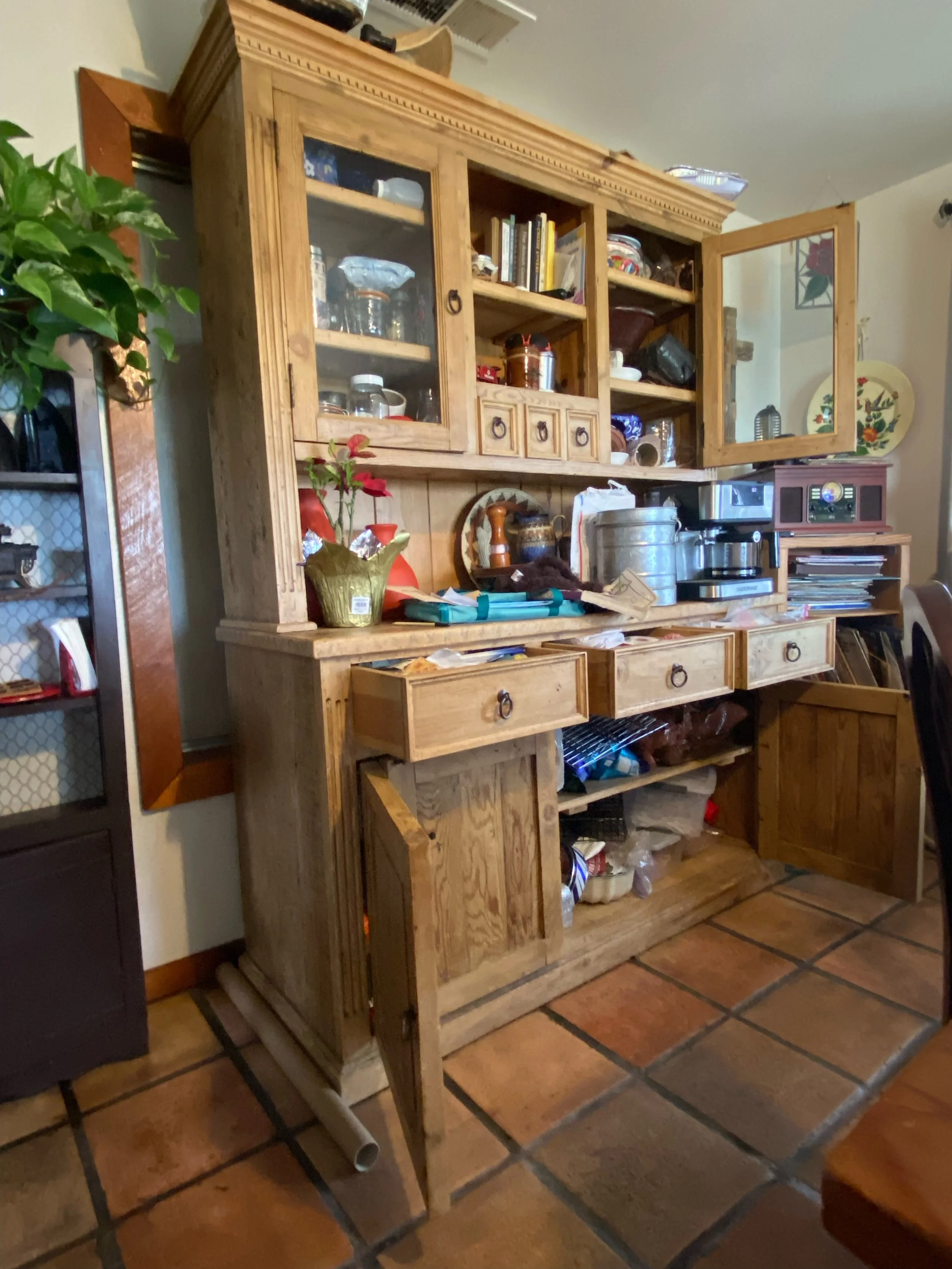 A wooden kitchen hutch with glass doors and drawers filled with dishes, cups, and kitchen items. The floor is tiled terracotta, and there is a potted plant to the left.