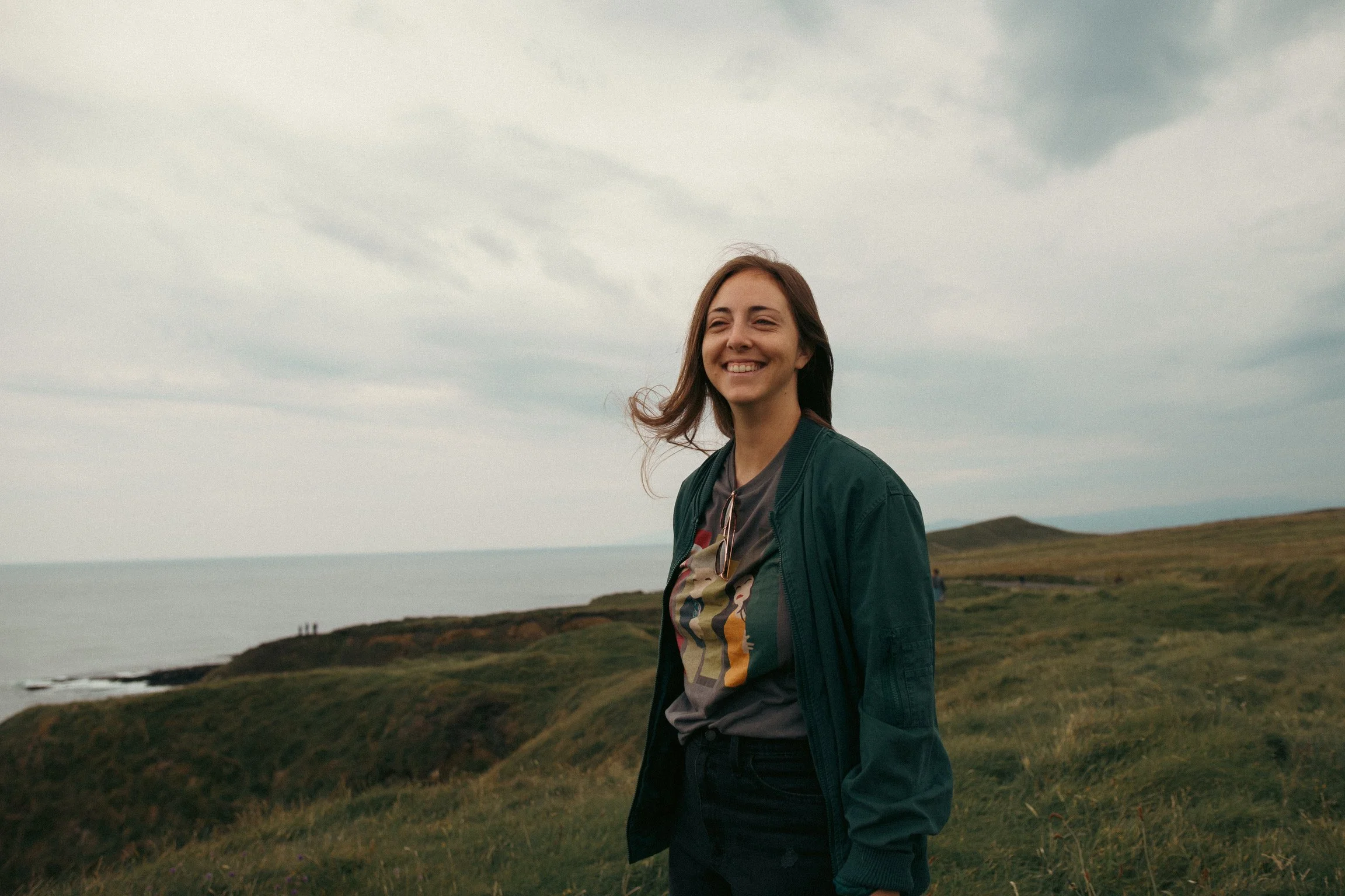 A young woman with brown hair smiling, standing outdoors on a cloudy day in a grassy landscape near the ocean.