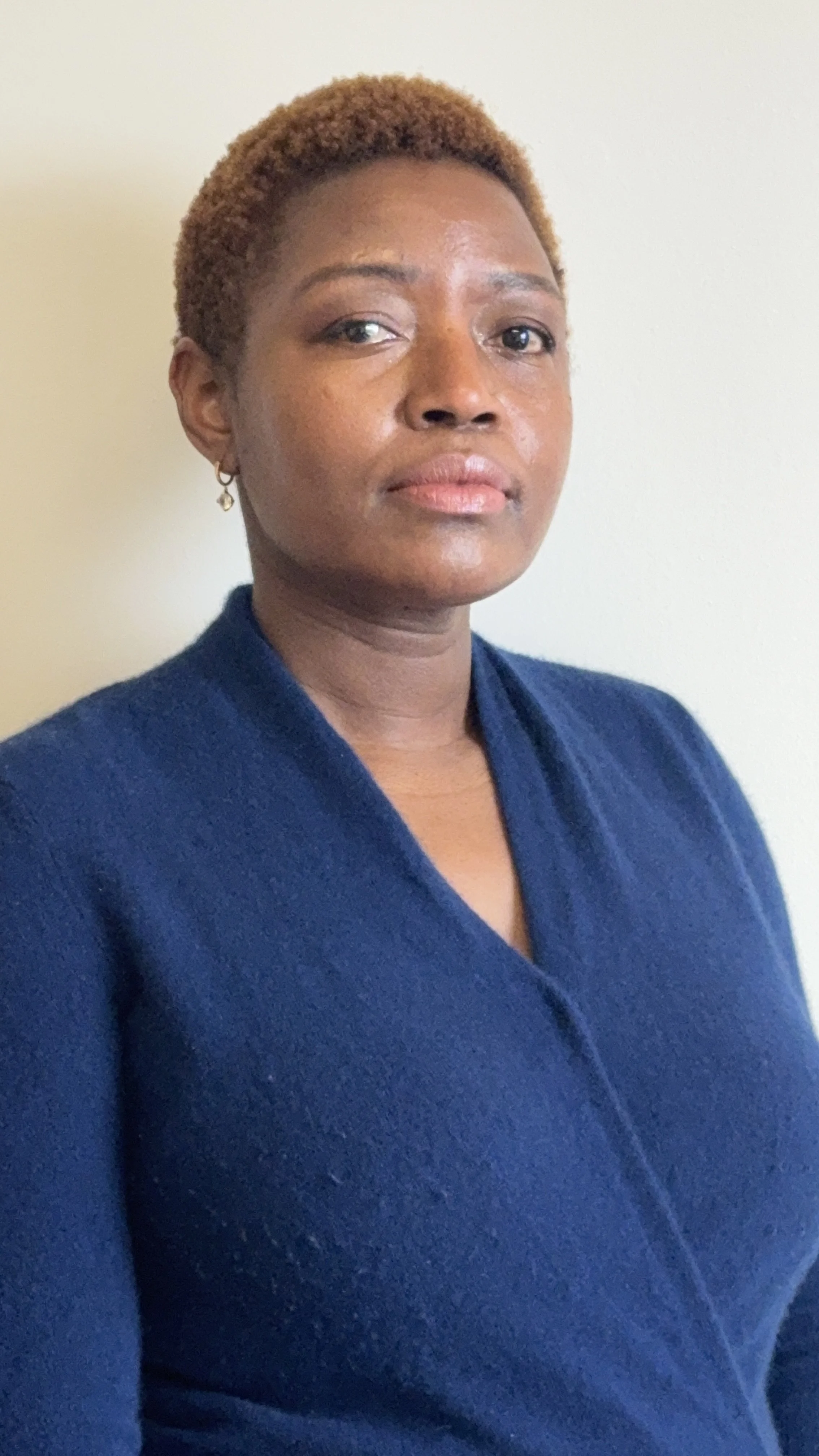 A woman with short, reddish-brown hair, wearing a dark blue blazer, standing against a light-colored wall, looking directly at the camera.