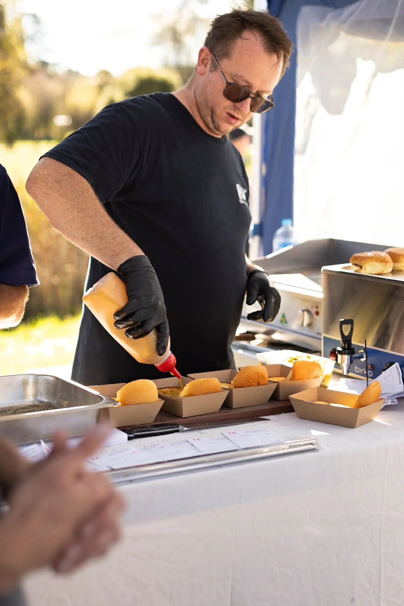 Man wearing black shirt, sunglasses, and black gloves preparing hot dogs with ketchup at an outdoor food stand during daytime.