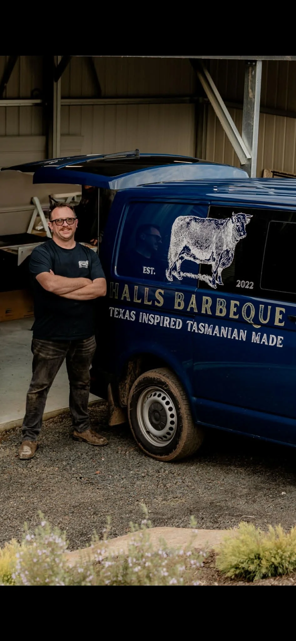 A man standing with crossed arms next to a blue van with a logo of a cow and the text "Halls Barbeque, Texas inspired Tasmanian made" in a garage or warehouse.