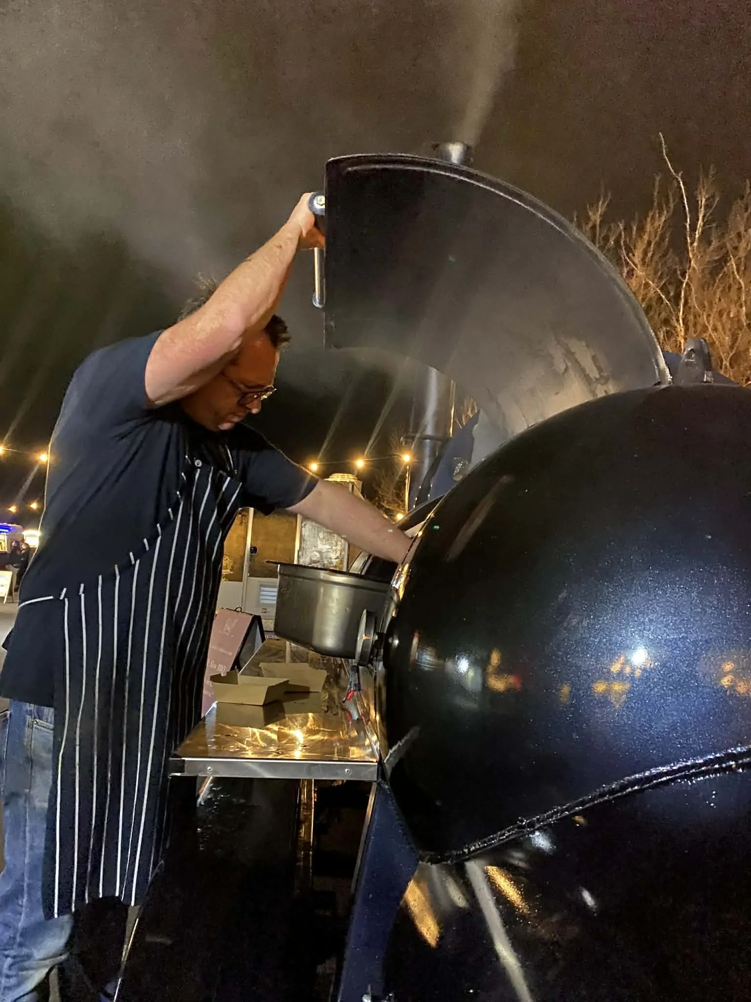Man in apron and glasses loading food into a large black barbecue smoker outdoors at night with string lights in the background.