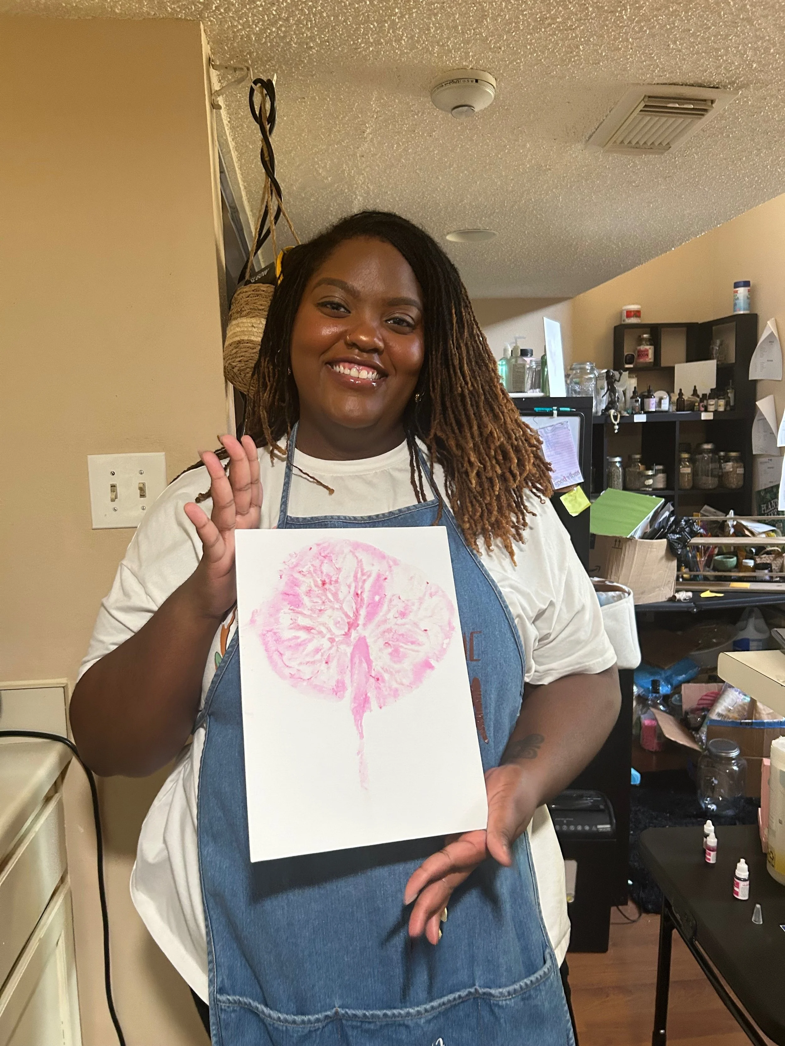 A woman with shoulder-length dreadlocks smiling and holding a watercolor painting of a tree with pink leaves in front of her.