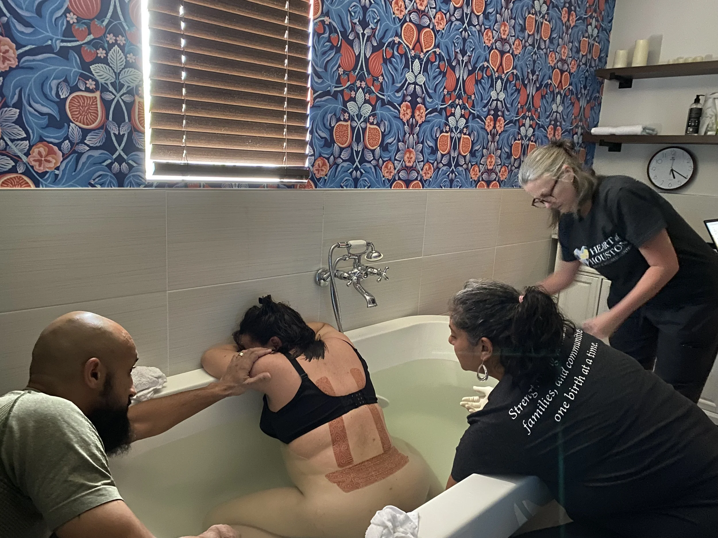 A woman is being baptized in a bathtub by three people, with two women supporting her and a man holding her head. The setting appears to be a room with floral wallpaper and a window with blinds.