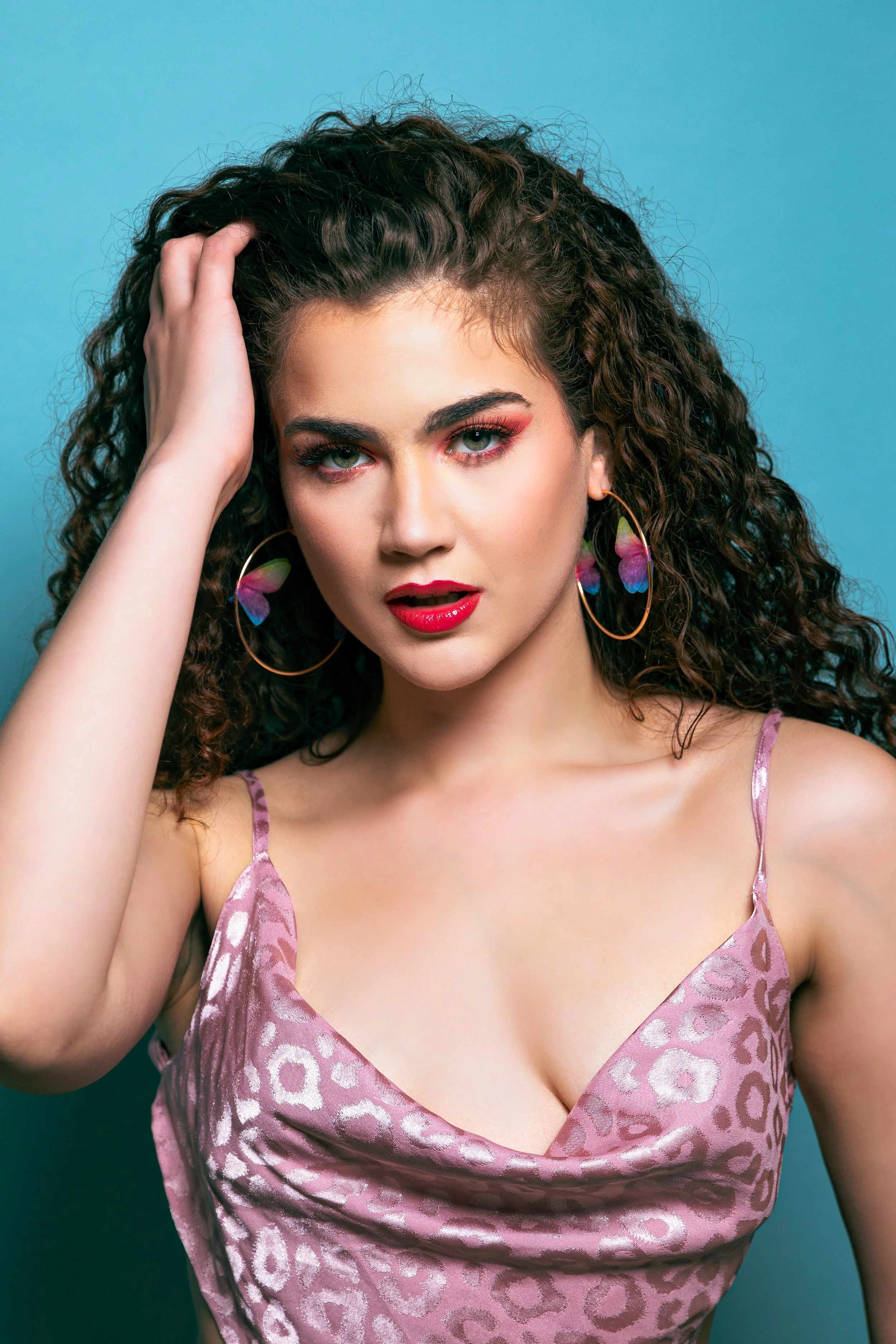 A woman with curly brown hair and striking makeup, wearing large colorful butterfly earrings and a pink leopard-print spaghetti strap top, posing with her hand on her head against a blue background.