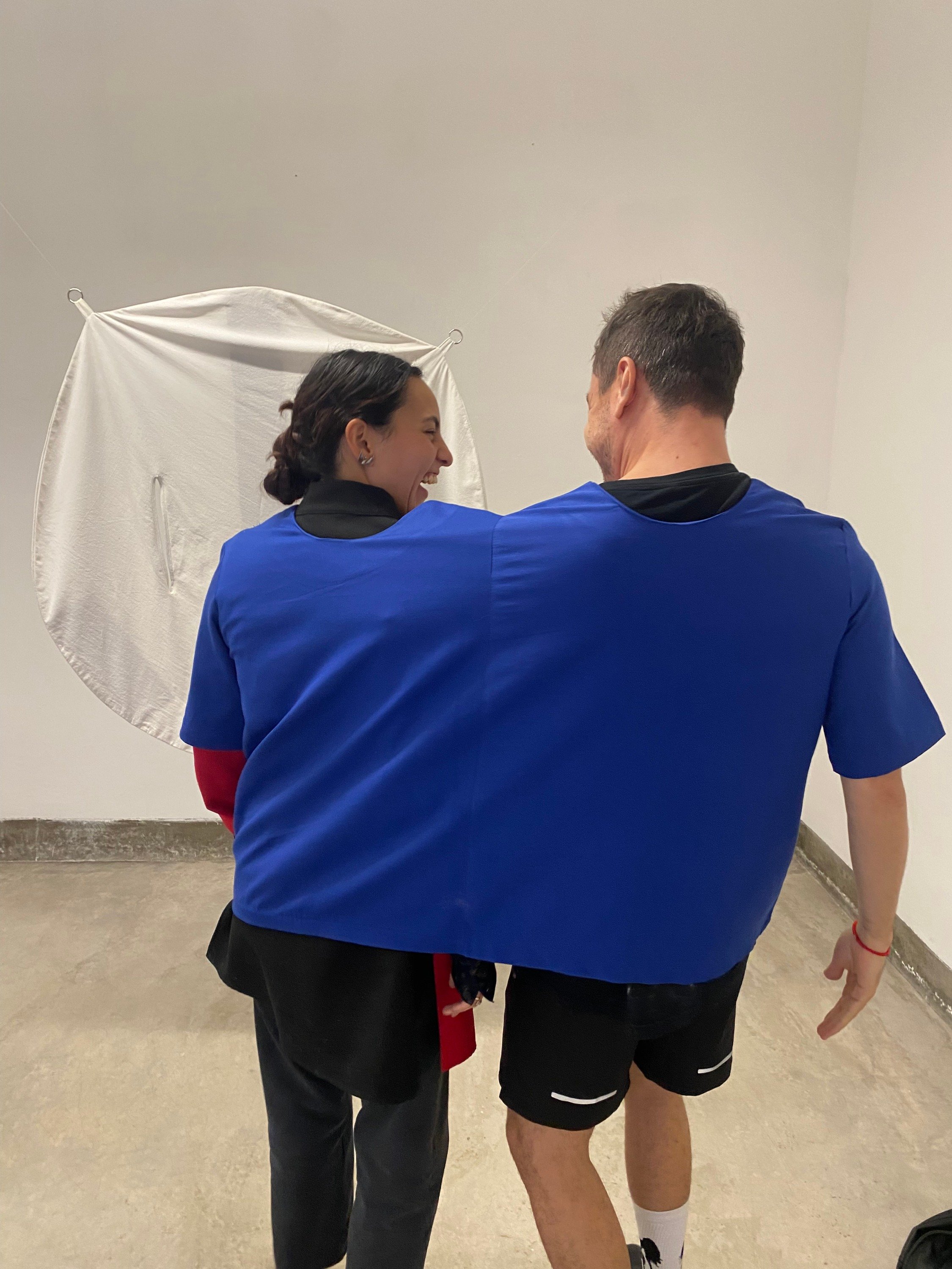 Two people, a woman and a man, are smiling and standing close together in a minimalistic room with plain white walls. They are wearing athletic clothing, with the woman in a black shirt with red sleeves and gray pants, and the man in a blue shirt and black shorts. There is a large, white umbrella reflector behind them.