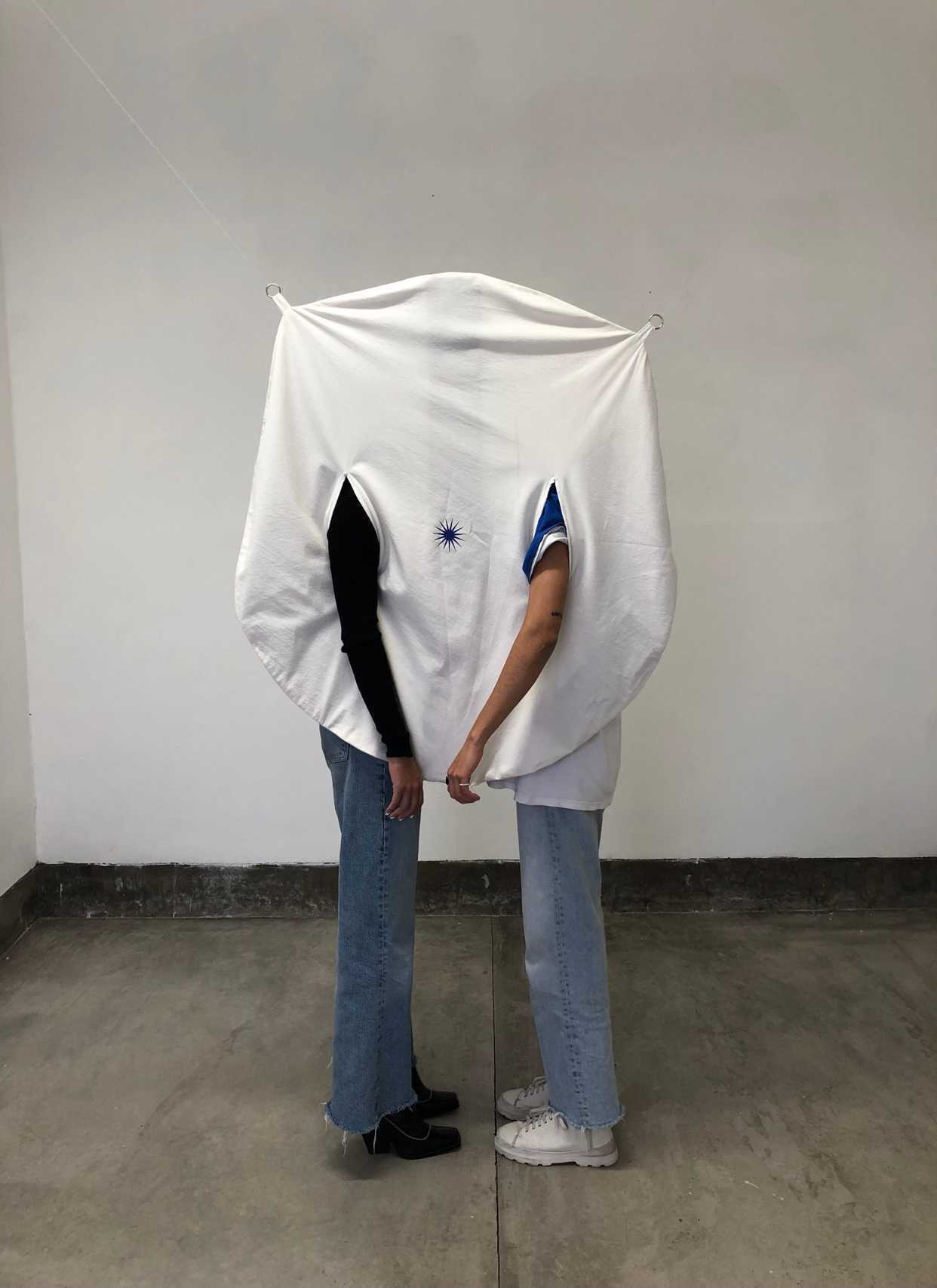 Two people standing close together, with their heads and upper bodies concealed by a large, circular white fabric with cutouts for the arms and a small blue star symbol in the center. Their lower bodies and legs are visible.
