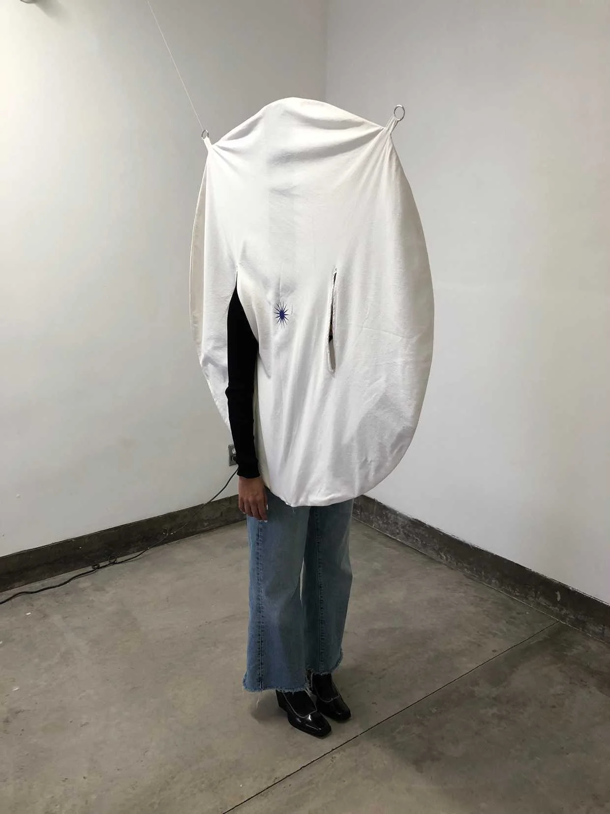 Person wearing a large, circular white fabric costume with cutouts, standing in a gallery or exhibition space.