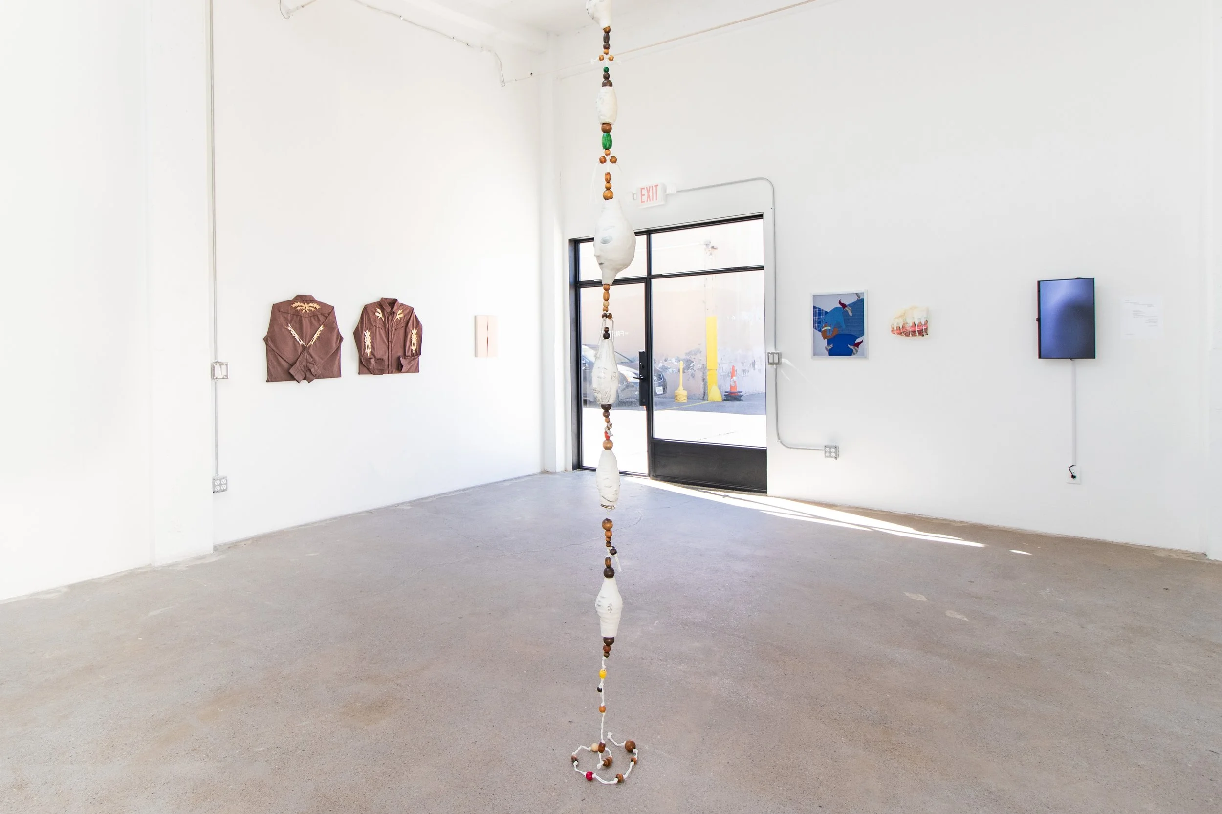 An art gallery with a hanging sculpture made of fabric and beads in the center, and framed artwork on the white walls. There is an exit door and a window showing the outside parking lot.