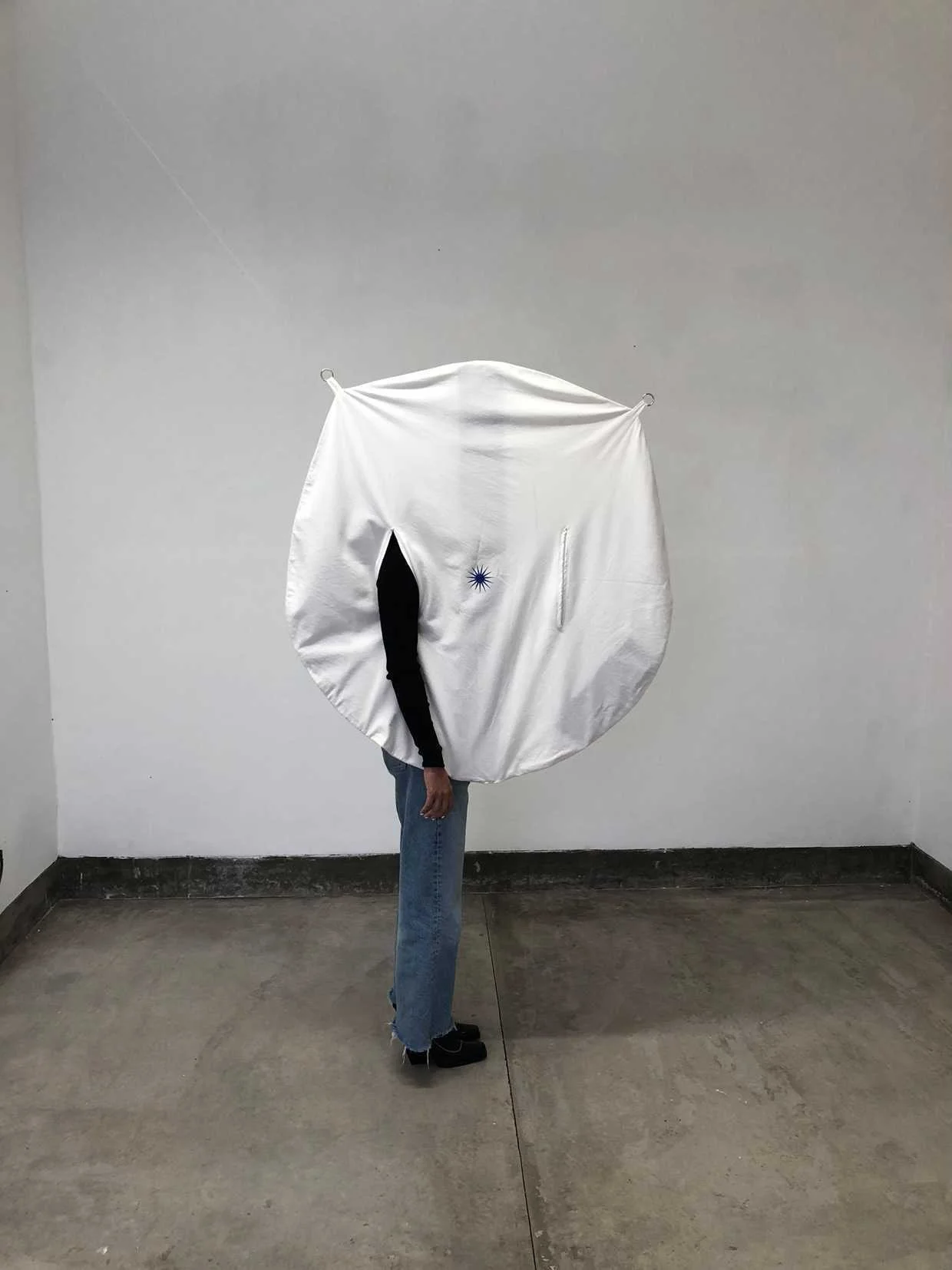 Person standing in a plain room with a large circular white fabric structure covering their upper body, with part of their arm and leg visible.