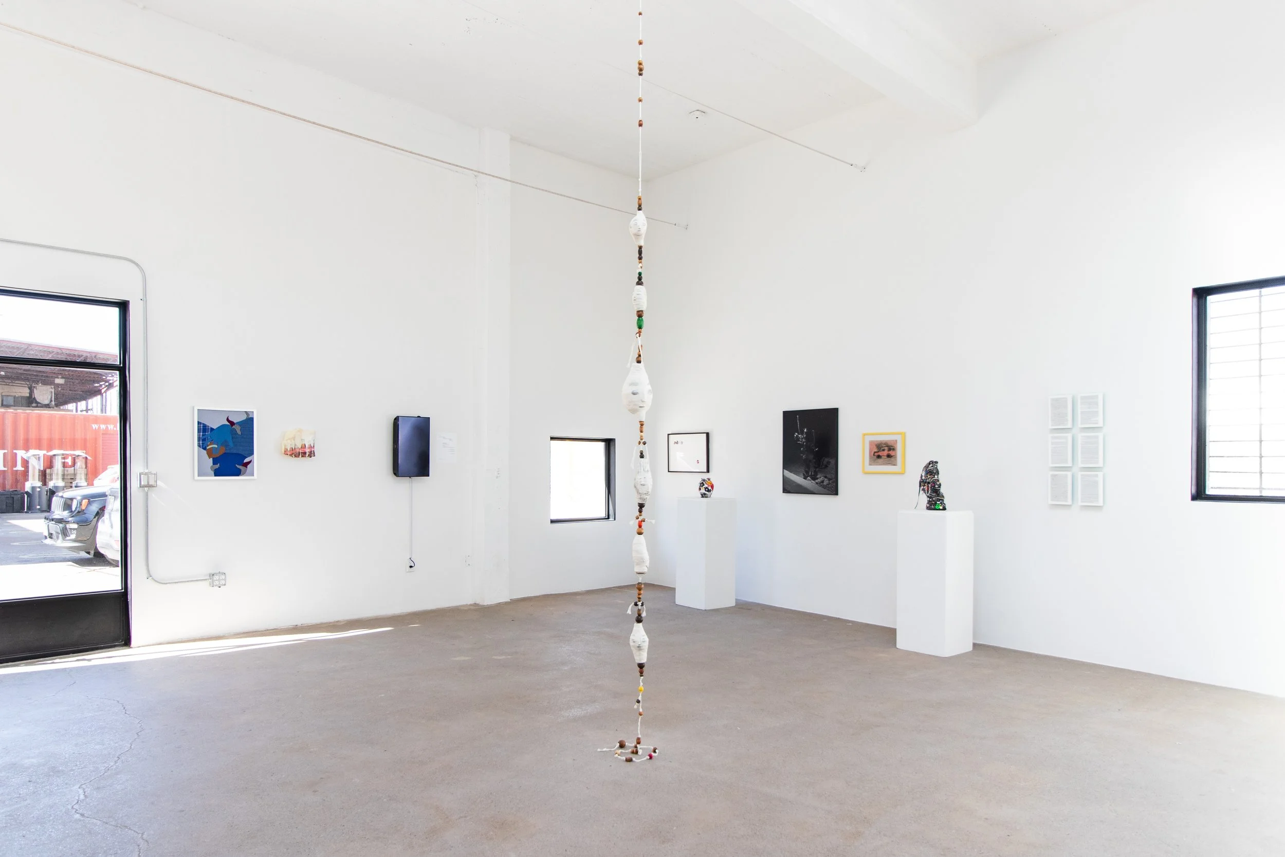 Modern art gallery with white walls and multiple art pieces on display, including paintings, photographs, and sculptures. A long beaded or ceramic hanging sculpture is suspended from the ceiling in the center of the room.