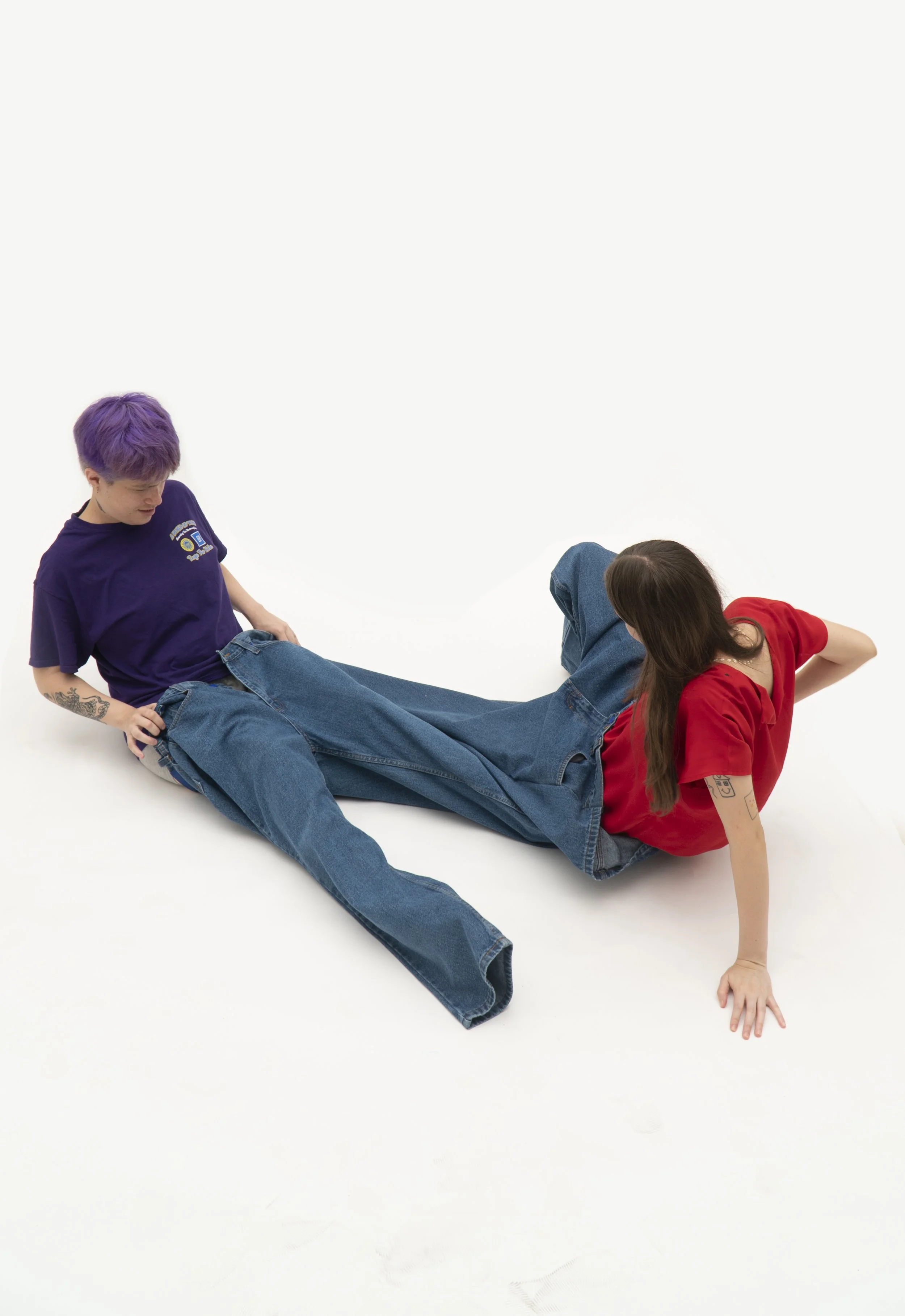 Two people are lying on a white surface, one with purple hair and a purple t-shirt, the other with brown hair and a red t-shirt, both wearing jeans. They are positioned facing each other, with one person on their back and the other on their side.