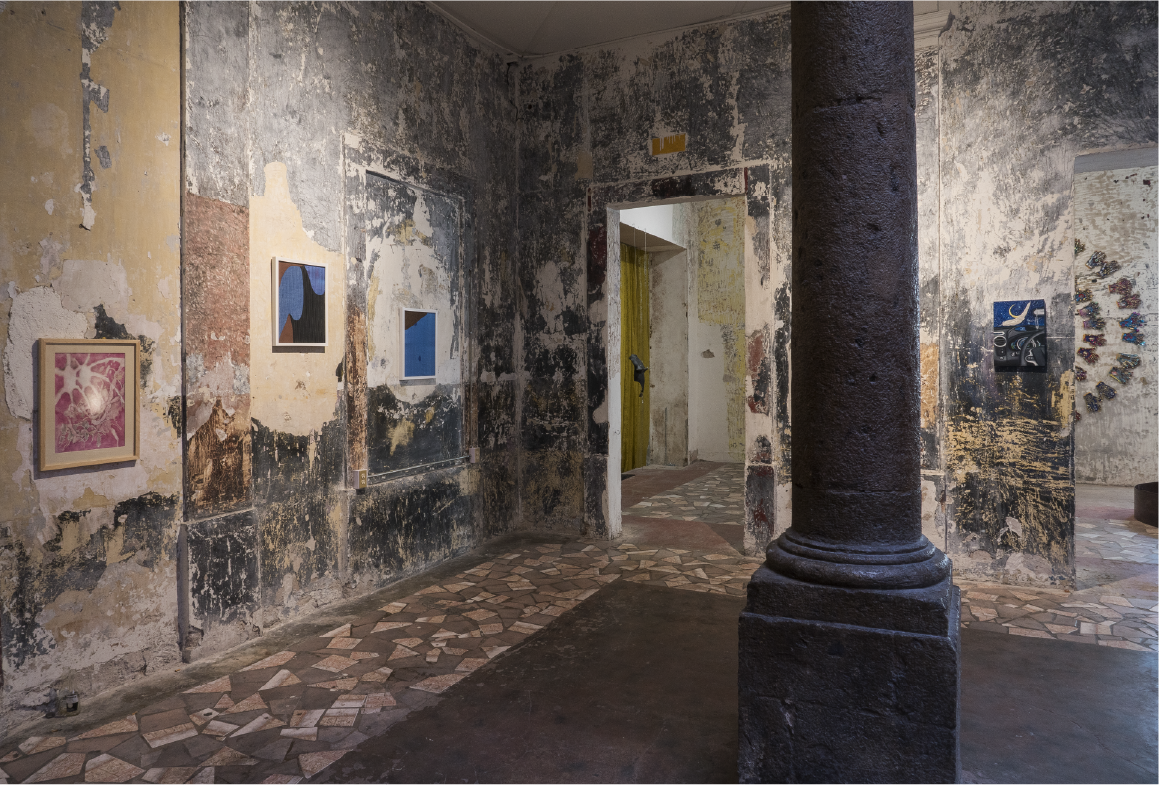 An art gallery with distressed, peeling walls and a stone column. Several colorful abstract paintings are displayed on the walls, and the floor has a patchwork of tiles. A doorway leads to another room with more artwork.