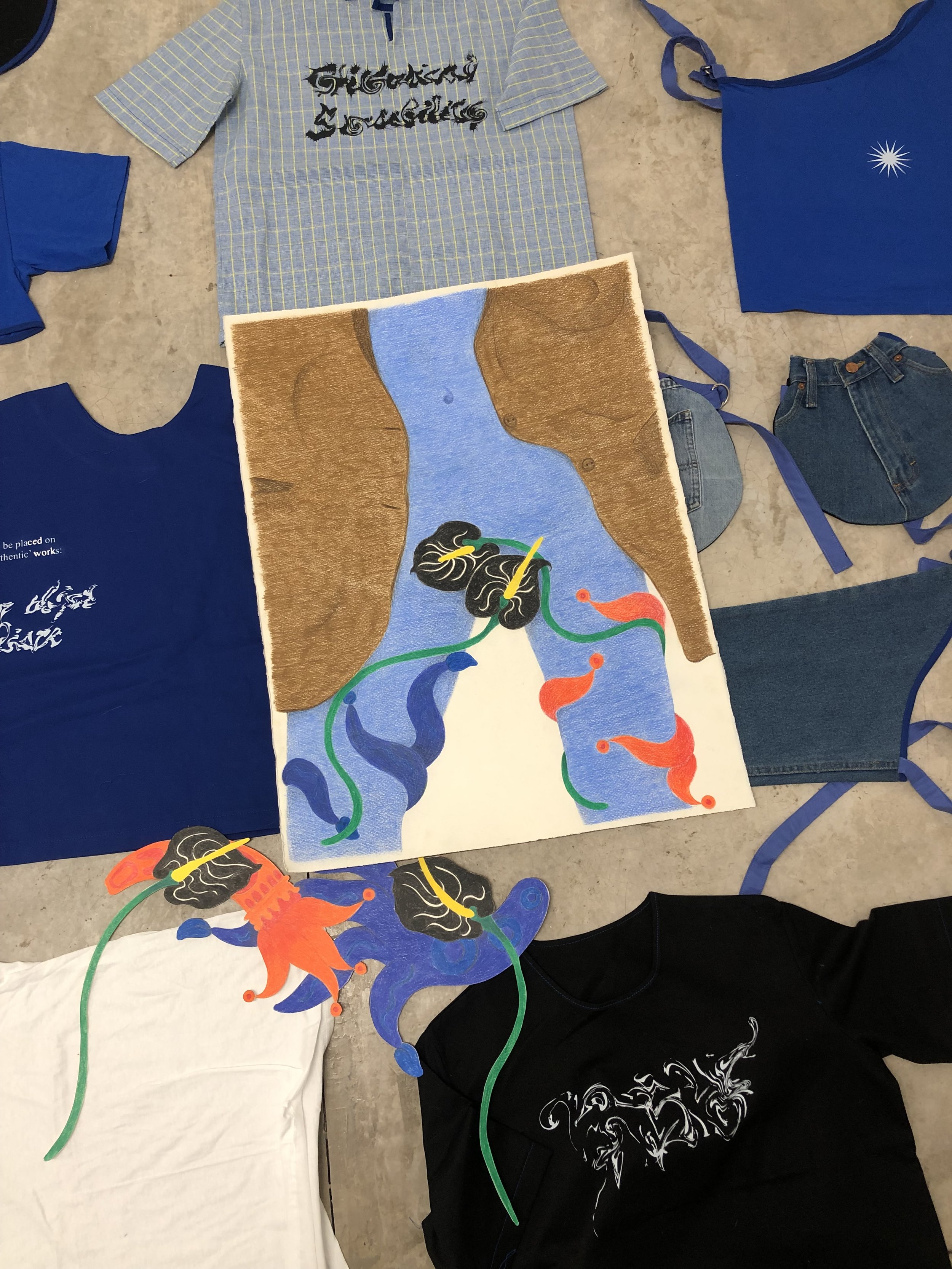 A collection of clothing and a colorful abstract artwork laid out on a concrete floor. The clothing includes a striped button-up shirt, a navy T-shirt, a white T-shirt, and denim shorts. The artwork features a surreal image with dark hair, a yellow beak, and colorful ribbon-like elements.