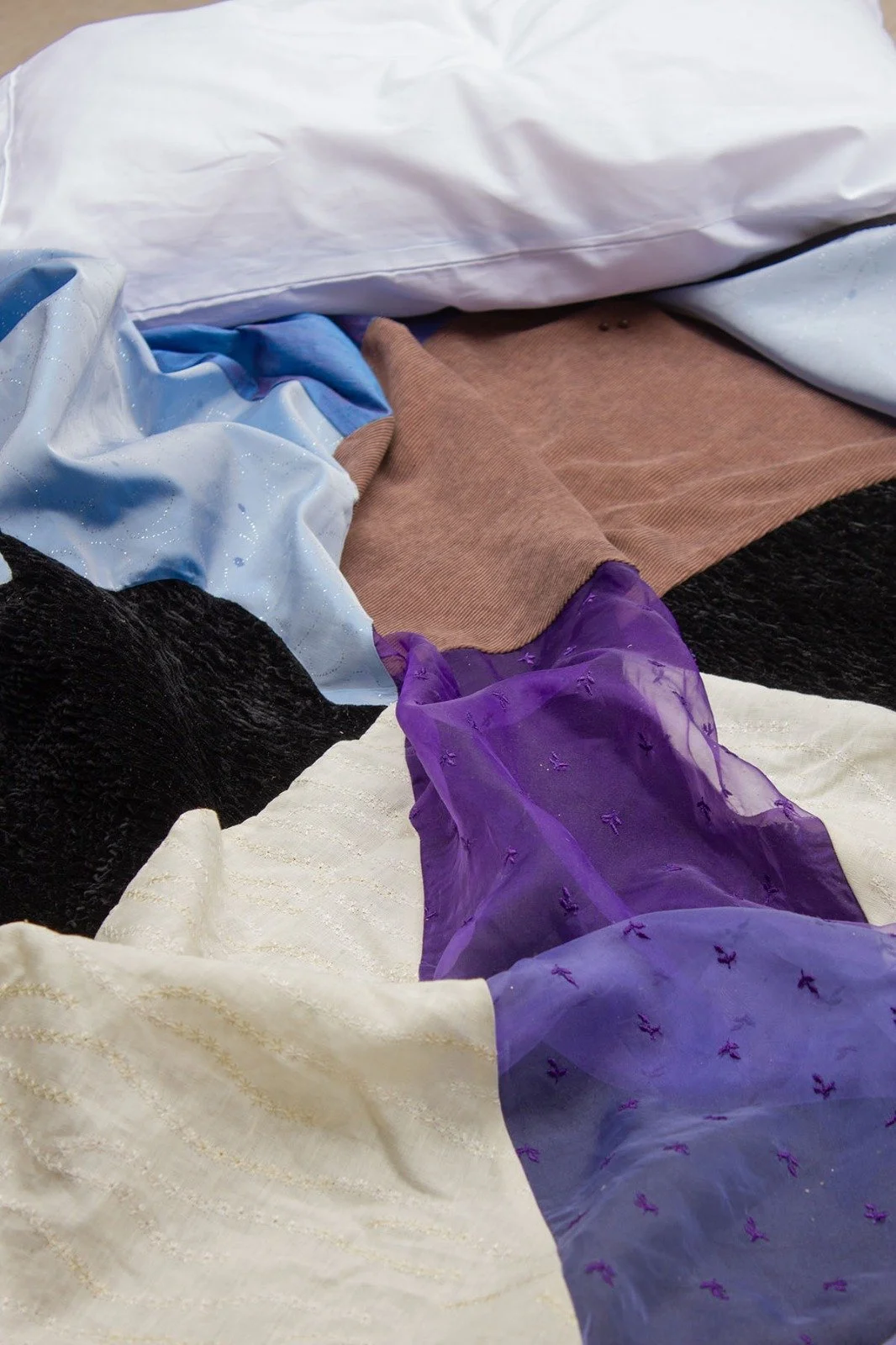 A bed with white bedsheets, brown pants, and various colorful fabrics, including purple, black, and light blue with glitter and embroidered details.