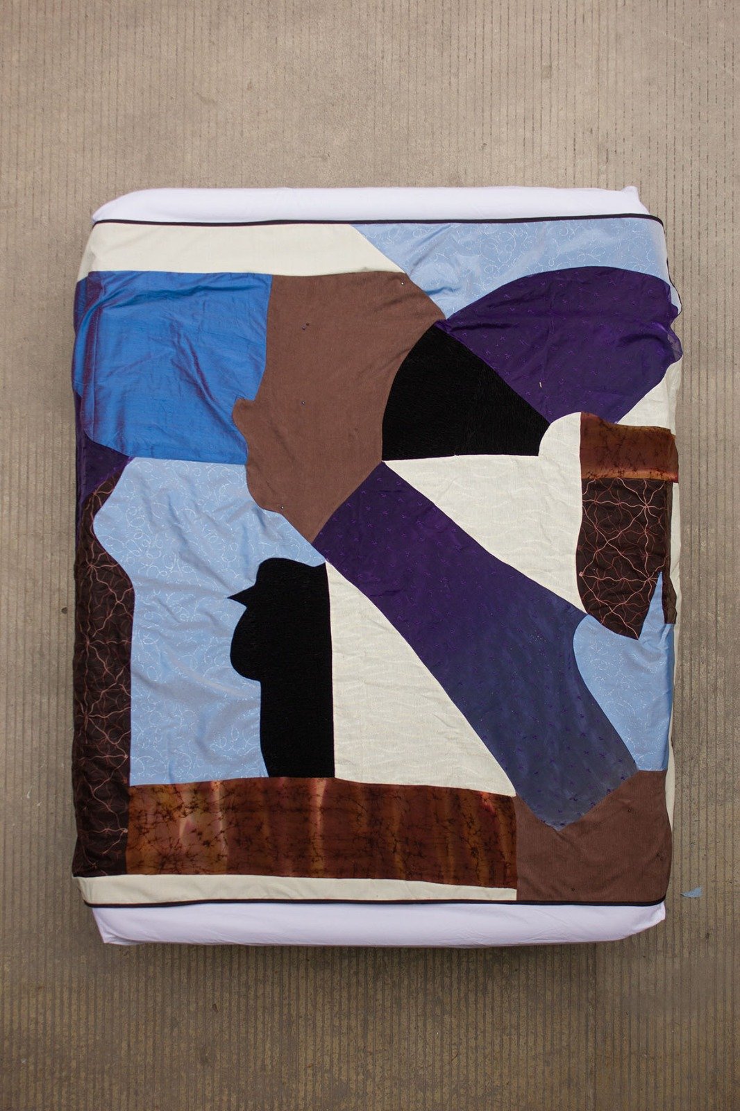 Colorful quilt with abstract shapes and a black cat silhouette, on top of a box on a gray surface.