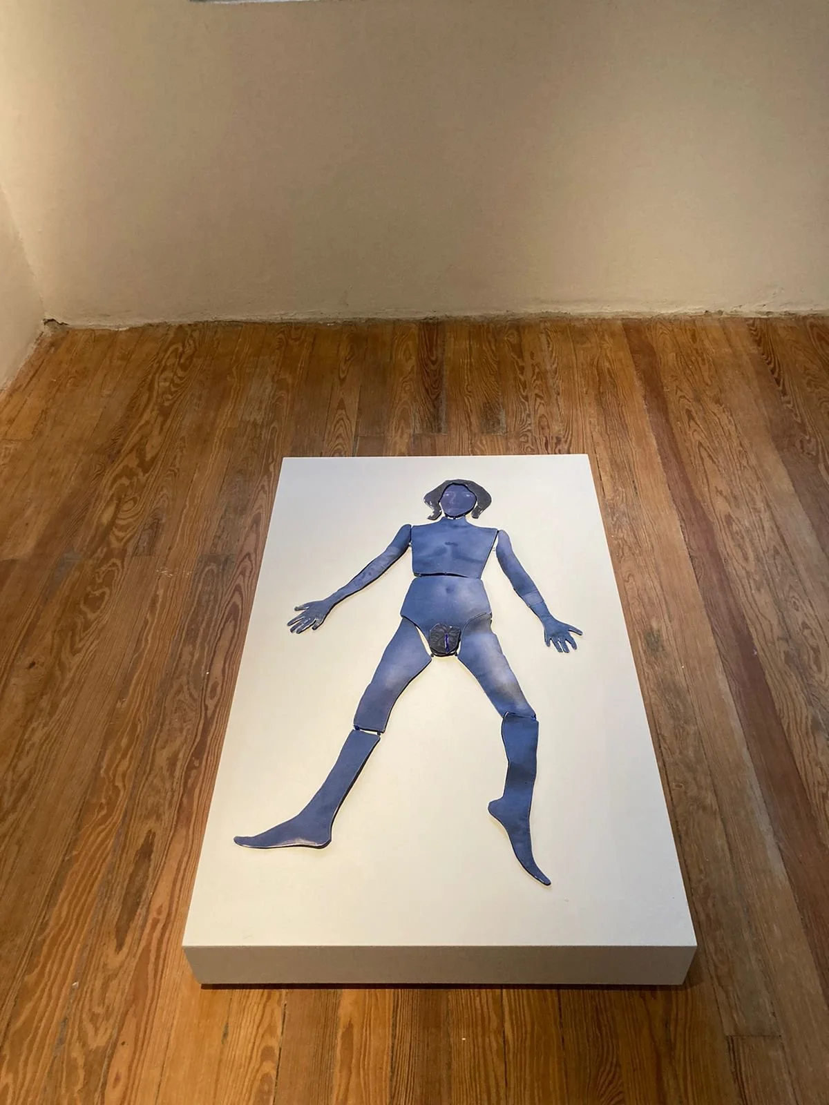 A piece of artwork on a white rectangular canvas depicts a stylized blue-toned figure of a person with the genital area cut out, laying flat on a wooden floor inside a room with plain beige walls.