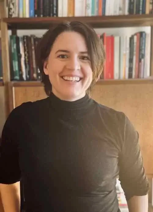 person with short brown hair wearing a turtleneck standing in front of a bookshelf