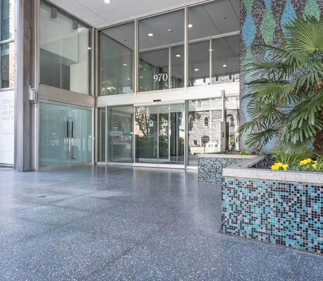 Entrance to a modern office building with glass doors, concrete flooring, and a tiled planter with green plants and yellow flowers.