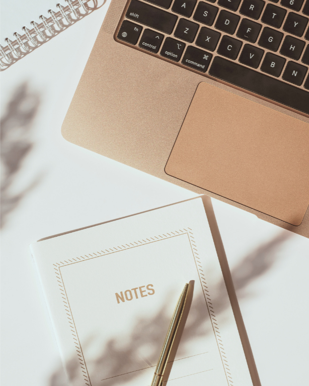 A close-up view of a beige laptop keyboard, a white notebook with "NOTES" printed on the cover, and a gold pen resting on the notebook, on a white surface.