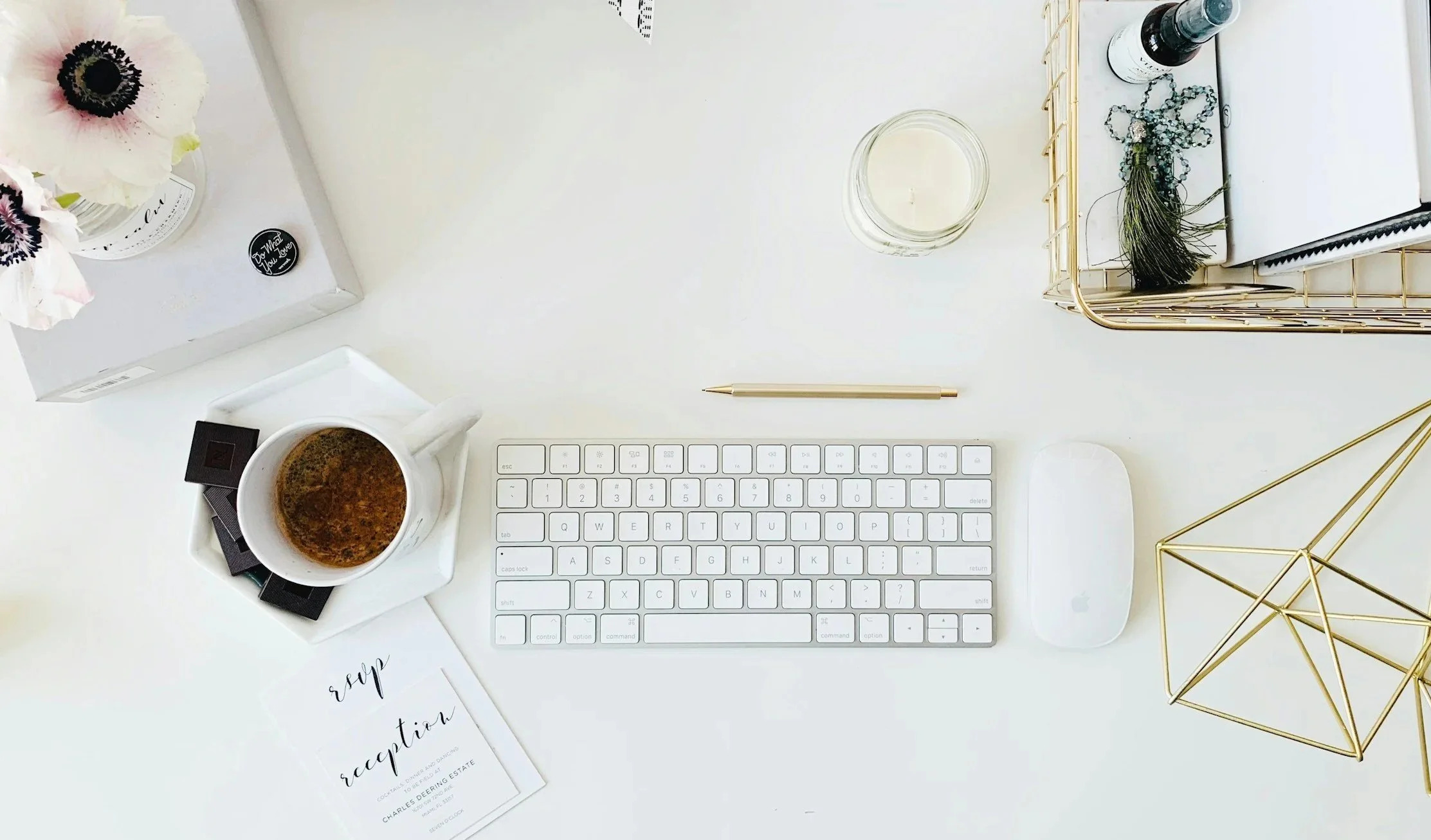 A clean, organized workspace with a white keyboard, a white computer mouse, a cup of coffee on a saucer, a gold geometric decorative object, a gold pen, a glass jar candle, a notebook with a pen, and a small floral arrangement with purple and white flowers.