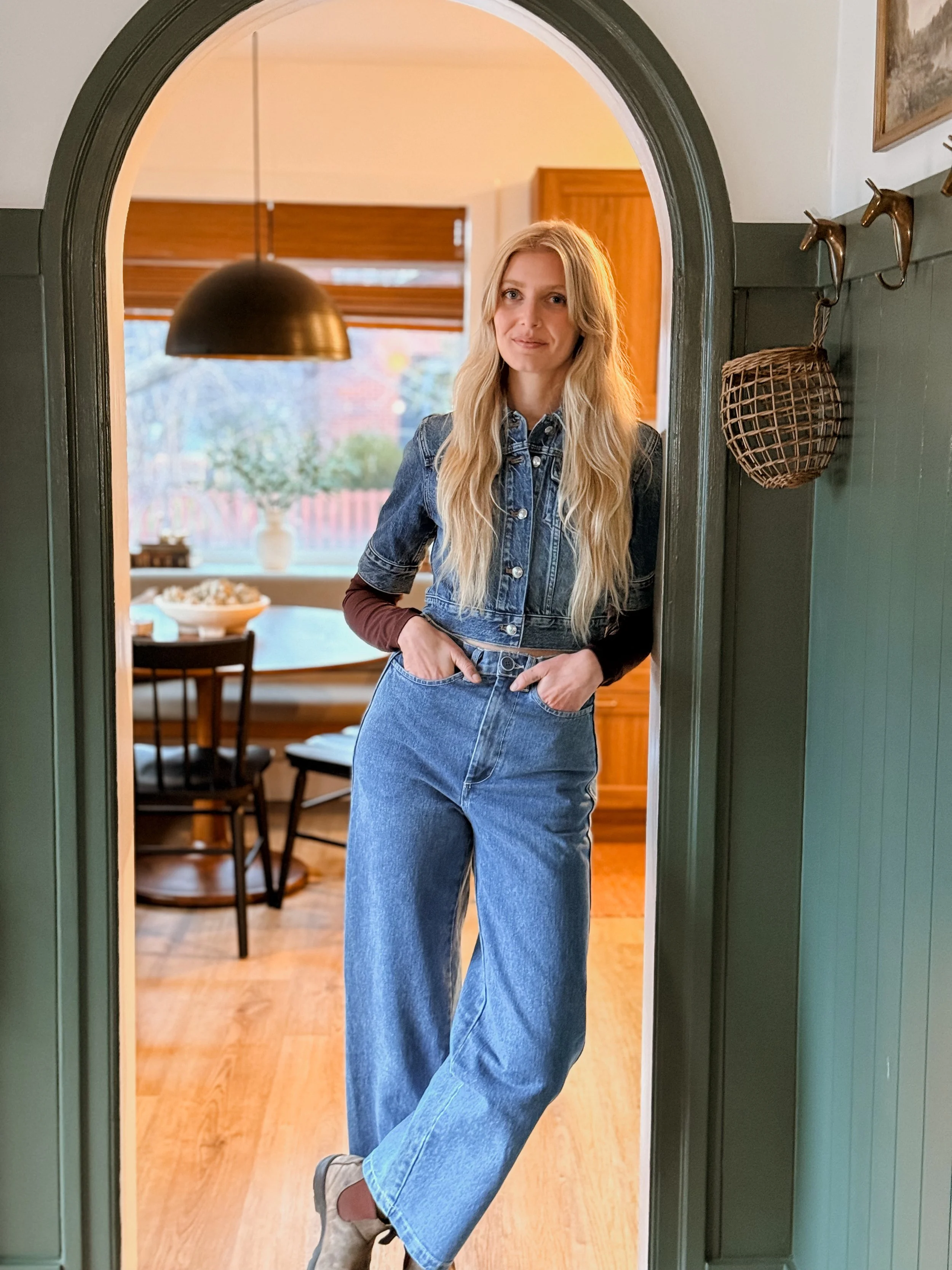 A woman with long blond hair standing in a doorway, wearing a denim jacket and jeans, with her hands in her pockets inside a cozy home.