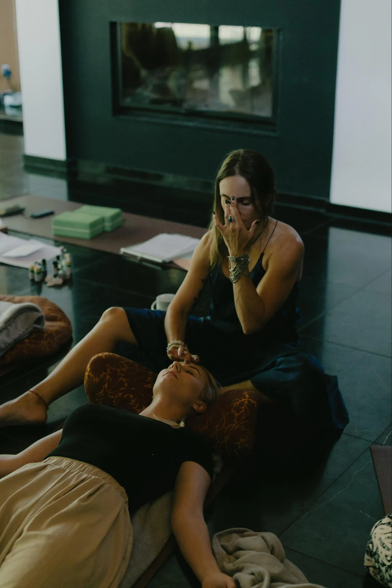 A woman receiving a Reiki healing session from another woman who is sitting on the floor with her hand over the woman's forehead in a room with dark flooring and a large fireplace.