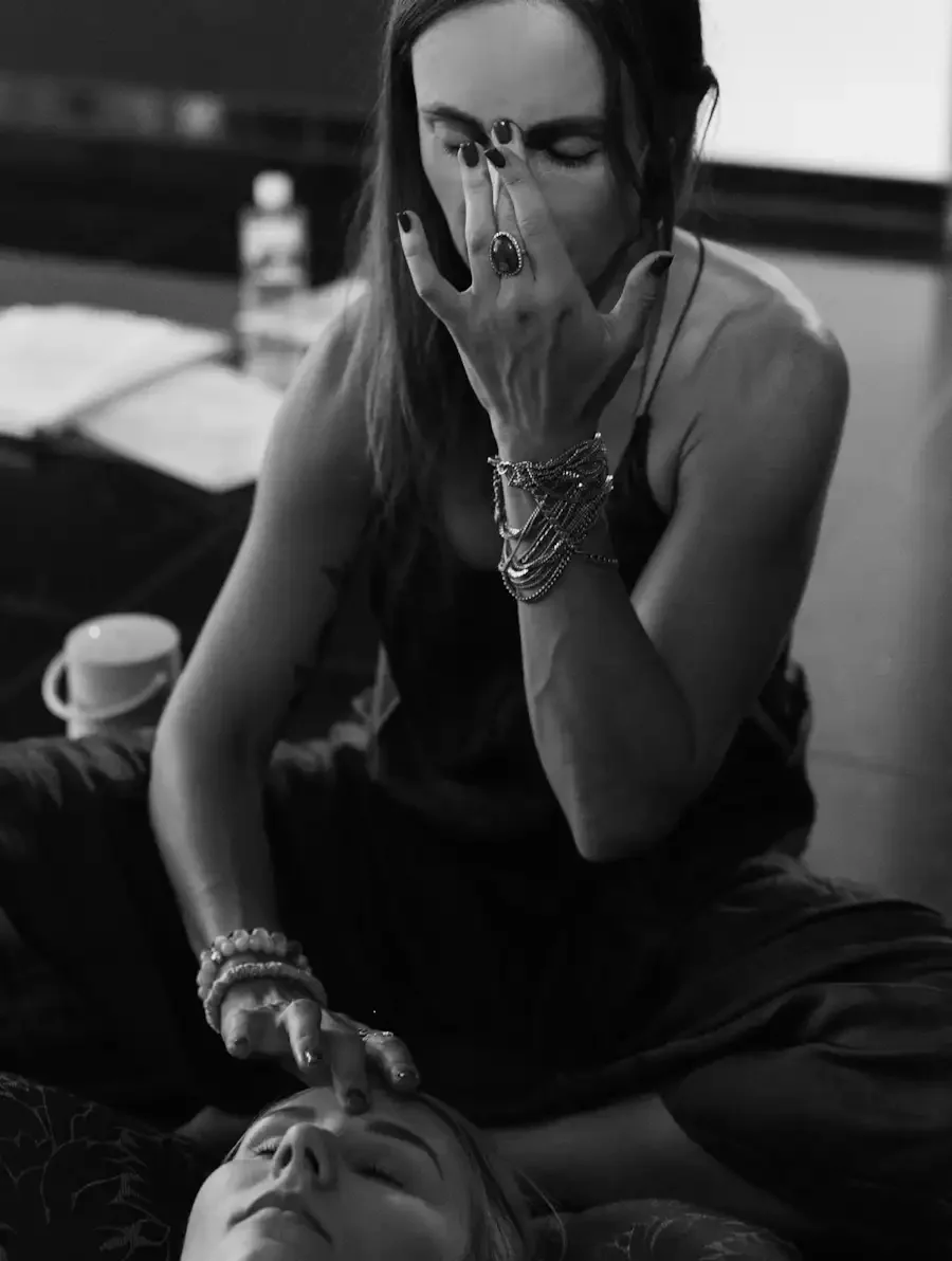 A woman with long hair and multiple bracelets, rings, and necklaces is transmitting a non-dual energy on a woman lying down with eyes closed.