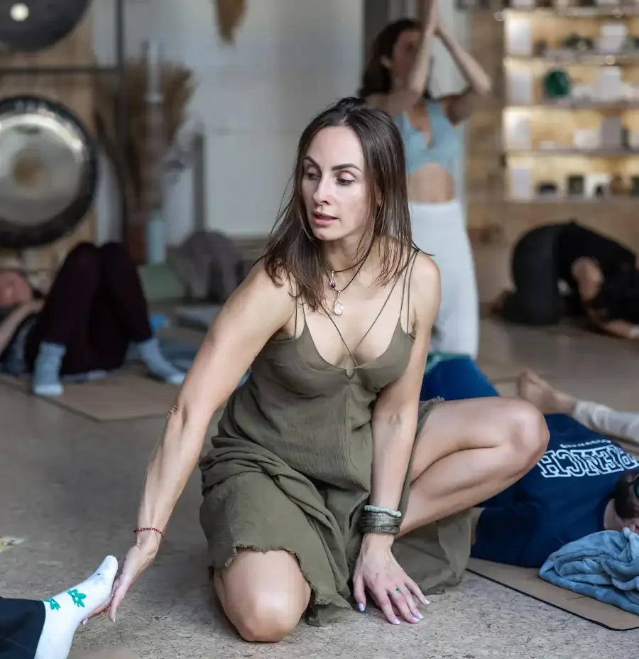 A woman practicing yoga or meditation in a studio with other people resting or practicing in the background.