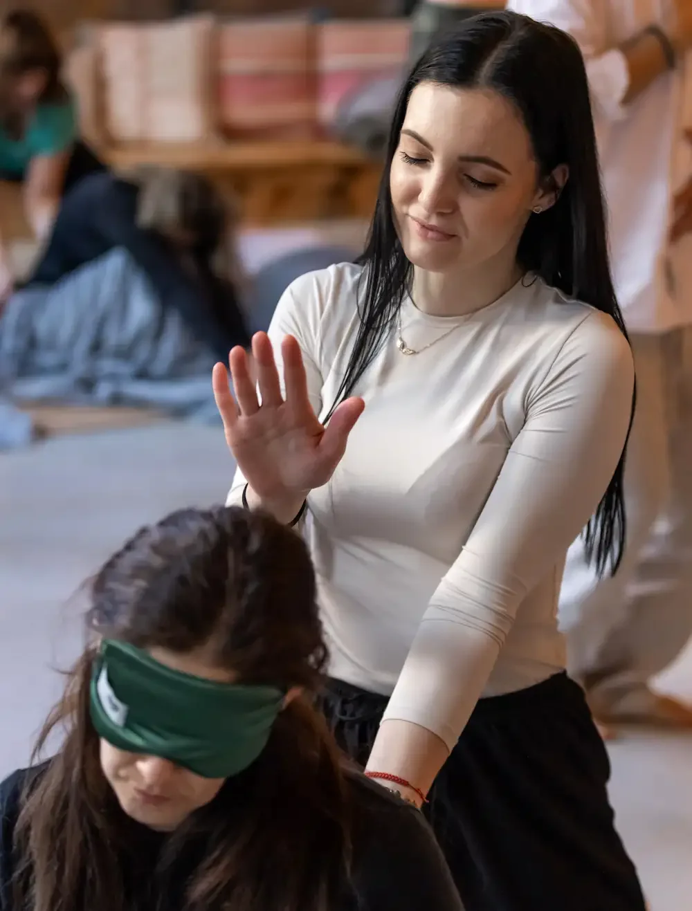 A woman with long black hair and light skin sends the energy out while standing behind a seated woman with brown hair, who is wearing a green eye mask; in the background, people are during a session.