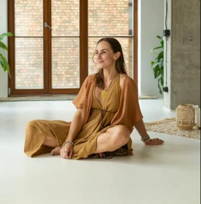 A woman sitting cross-legged on the floor inside a room with large windows and brick exterior visible outside. She is wearing a brown dress and accessorized with jewelry, smiling softly.