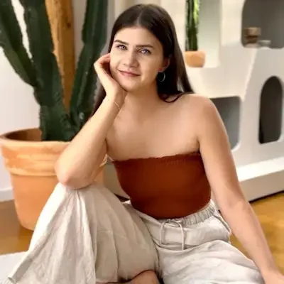 A woman with long dark hair, wearing a strapless brown top and light-colored pants, sitting on the floor inside a room with a large potted cactus, a white wall, and decorative shelves.