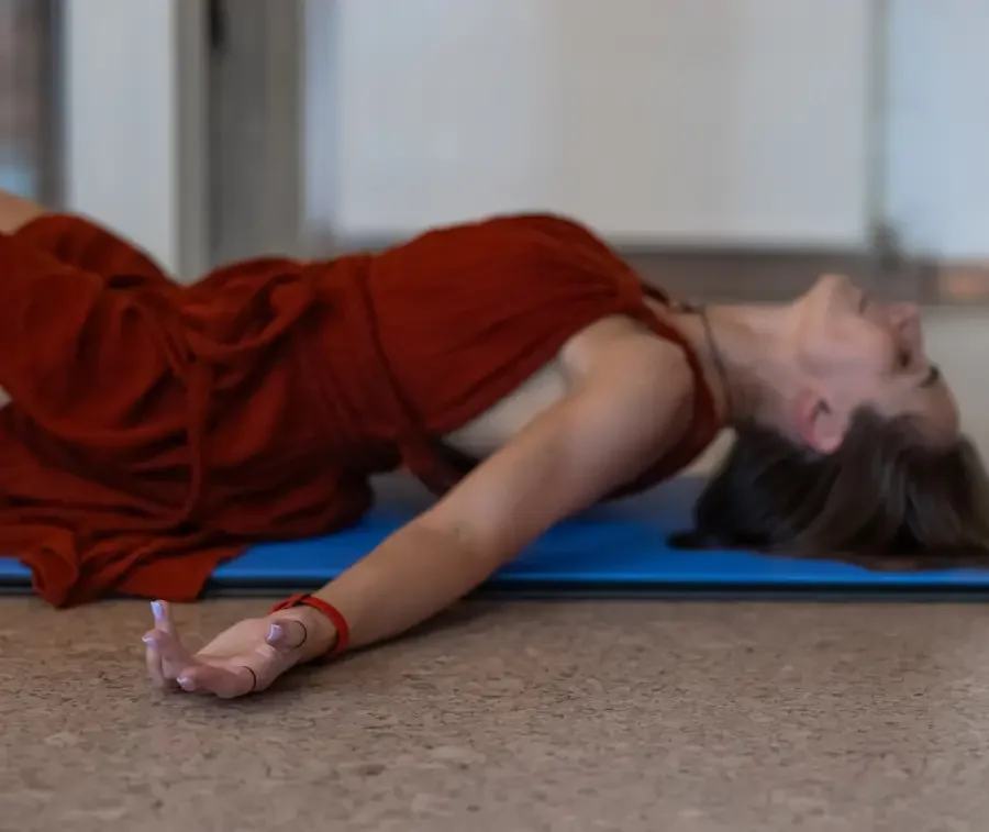A woman with long brown hair in a red dress lying on a blue yoga mat on the floor, stretching with one arm extended forward and face turned upward.
