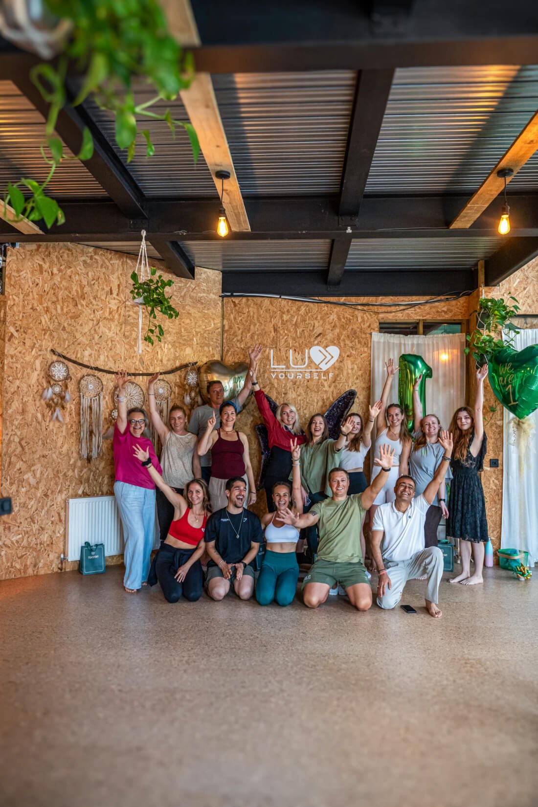 A group of people celebrating indoors with palms and peace signs, in front of a wooden wall decorated with dreamcatchers, greenery, and balloons, including a large gold heart and a green number 1 balloon, with a sign that reads 'Luv Yourself'.