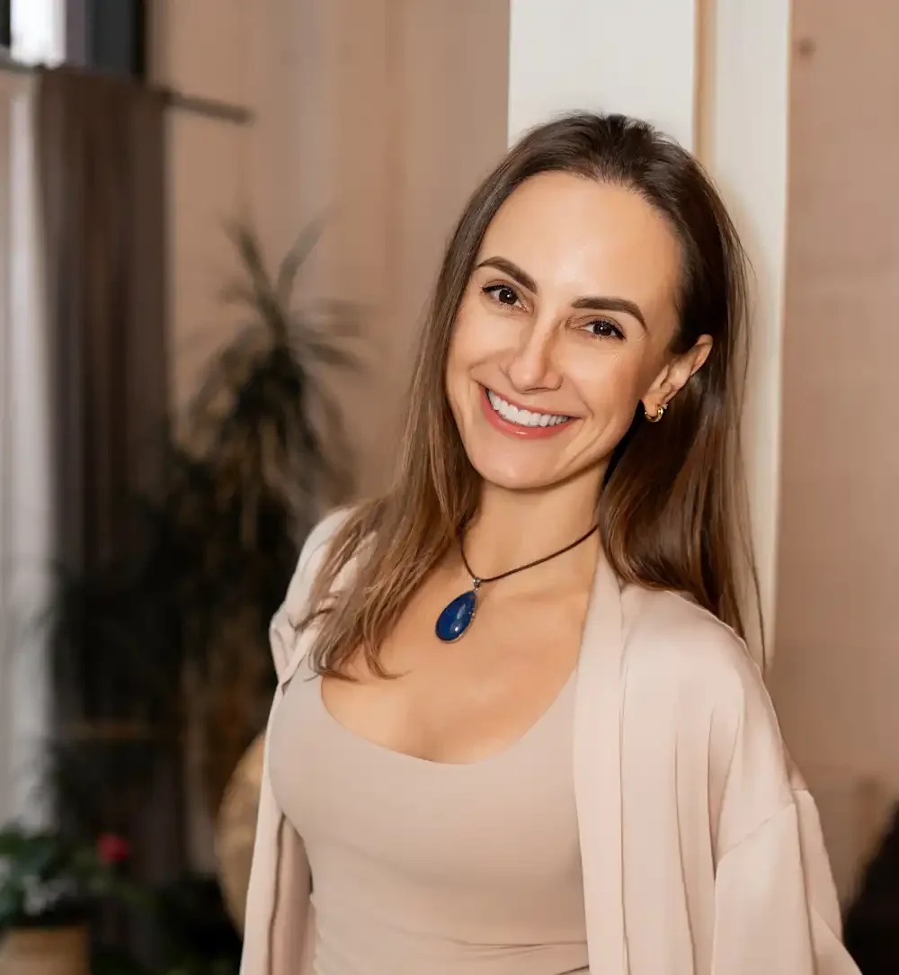 A smiling woman with brown hair wearing a beige outfit and a blue pendant necklace, standing indoors with a blurred background.
