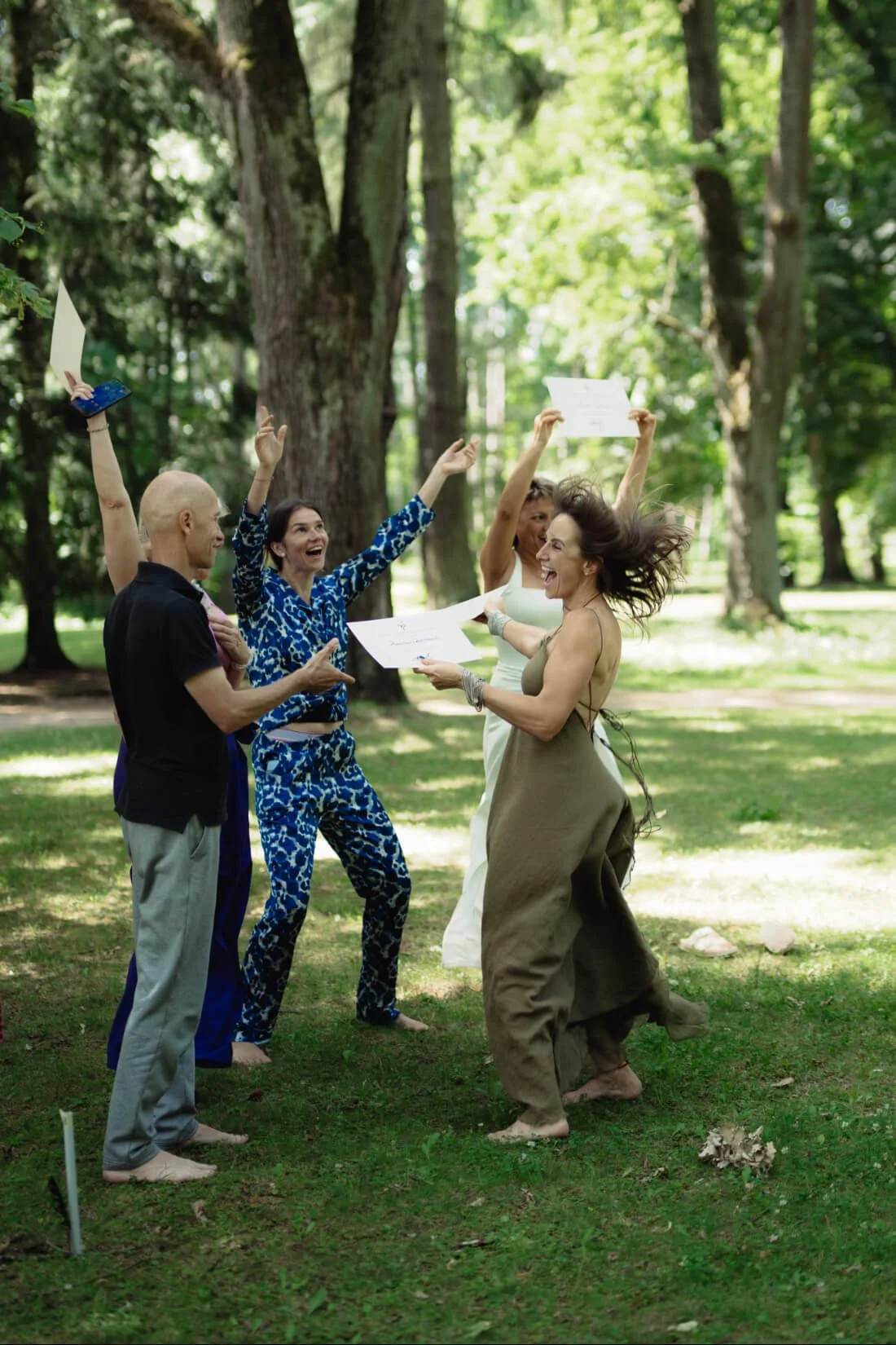A cheerful group of five women and one man outdoors in a nature, celebrating and holding certificates, with some raising their hands and smiling.