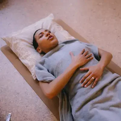 A woman on the cork flooring under the blanket holding her hands on belly and chest and exhaling with a peaceful face and eyes closed.